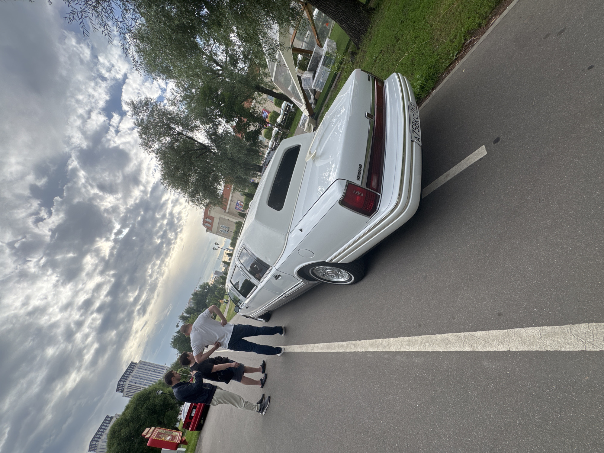 Three men stand on an asphalt road next to a white limousine. The men are facing away from the camera, and two of them are looking at the limousine. The man on the far left is wearing a dark jacket and light pants, the man in the middle is wearing a black t-shirt and shorts, and the man on the right is wearing a white t-shirt and dark pants. The limousine is a Lincoln Town Car, and it has a white vinyl top. The road is marked with a white line. In the background, there are trees, buildings, and a cloudy sky. The image was taken in Moscow, Russia, at an outdoor event. The architectural style of the buildings and the license plate on the limousine suggest this is Russia.