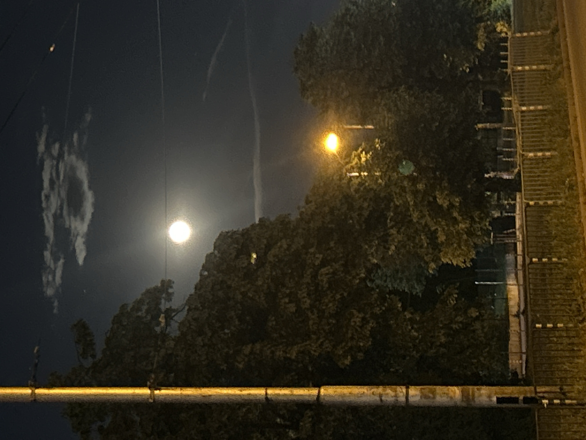 The image shows a night scene, likely in Sofia, Bulgaria. A bright, full moon dominates the left side of the frame, partially obscured by sparse clouds. To the right of the moon, a street lamp illuminates a cluster of trees. In the foreground, a weathered, possibly metallic, horizontal bar runs across the lower portion of the image. On the right side, a textured vertical surface is visible, possibly a wall or part of a structure. Thin lines, possibly electrical wires, stretch across the sky. A faint vapor trail can be seen in the upper part of the image. The lighting suggests an urban environment, but specific details of the location within Sofia are unclear. No people are visible.