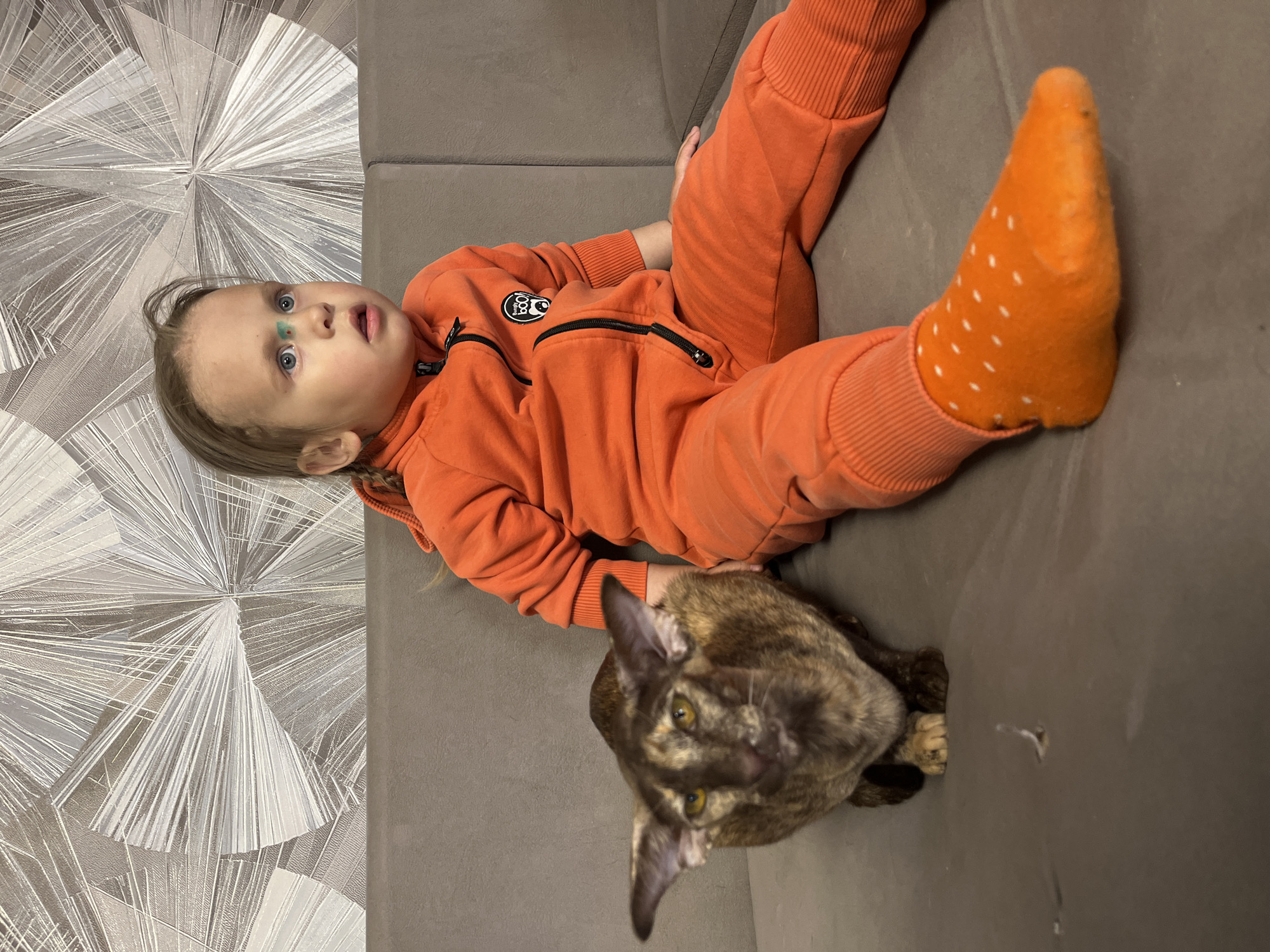 This indoor image, taken in Chaikovsky, Russia, features a young child and a cat on a grey sofa. A toddler, with light hair, is lying on their back or side, wearing a bright orange zippered jumpsuit. One visible foot is covered by an orange sock with small white polka dots. The child has blue eyes and a slightly open mouth, looking upwards and to their right with an expression of curiosity or mild surprise. A small green mark is present on the bridge of the child's nose.
Next to the child, a domestic cat with a distinctive tortoiseshell or tabby coat pattern and luminous yellow-green eyes is positioned. The cat's head is slightly tilted as it looks directly towards the camera, appearing calm and observant.
The sofa is a plain grey fabric. Behind the subjects, a wall is adorned with modern, geometric wallpaper in shades of grey and white, characterized by radiating lines and abstract, fan-like patterns. On the child's orange jumpsuit, there is a circular black patch featuring a white cartoon character or logo, which appears to include indistinct white text or numbers, possibly "100". Black zippers are also visible on the jumpsuit. The scene is illuminated by indoor lighting.