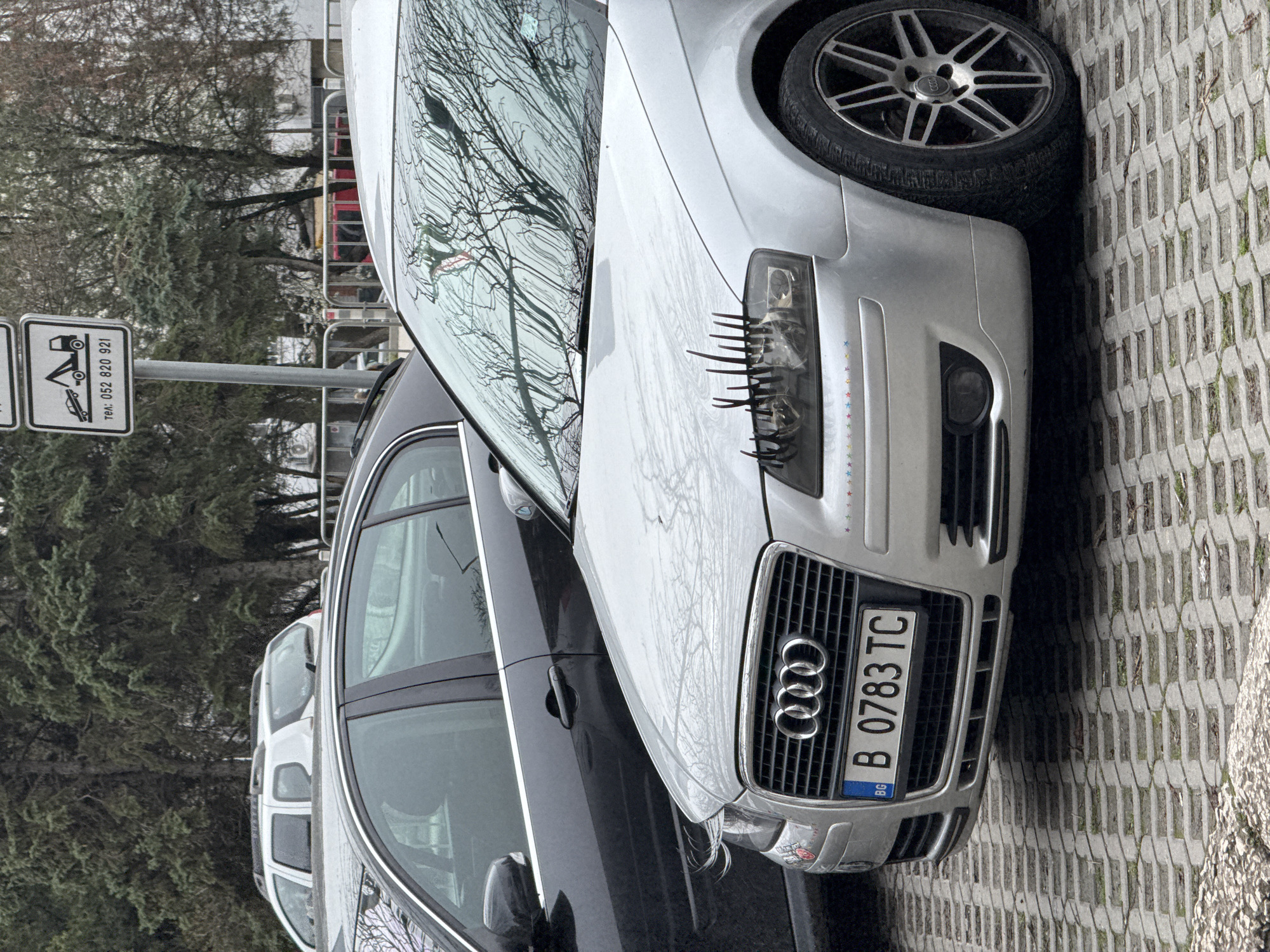 The image shows a silver Audi A3 parked on a paved surface in Varna, Bulgaria. The car has eyelashes attached to the right headlight. The license plate reads "B 0783 TC". To the left of the Audi, there's a black car and a street sign depicting a tow truck symbol with text including "тел. 052 820 921." The background consists of trees and a metal fence. The sky appears overcast.