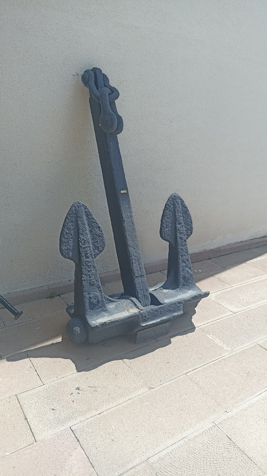 An old, heavy, black metal anchor is leaning against a textured, light-colored wall. The anchor has a long, thick shank with a shackle at the top and two flukes at the bottom, each with a pointed, leaf-like shape. The anchor rests on a light-colored, paved ground made of rectangular stones. The scene appears to be outdoors, possibly in a public space, as indicated by the paving and the wall. The lighting suggests it is daytime, with shadows cast on the ground. There are no people visible in the image. The anchor is positioned slightly to the right of the center of the frame. A small, metallic label is affixed to the shank of the anchor.