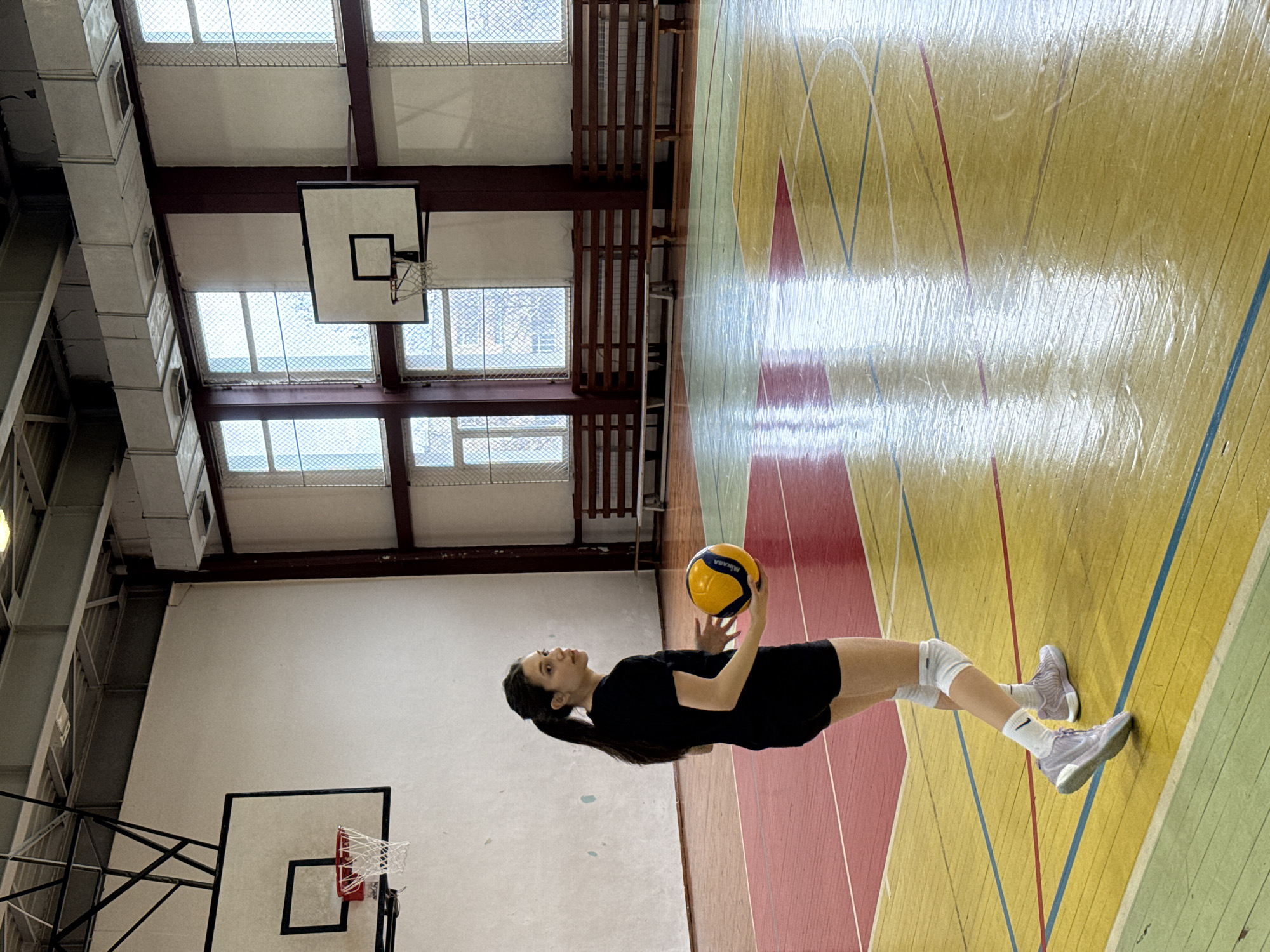 A young woman in a black t-shirt and black shorts stands on a gymnasium floor holding a yellow and blue volleyball. She is wearing white athletic shoes and knee pads. The gymnasium has a wooden floor with painted lines, and a basketball hoop is visible on the wall. The gym is located indoors with a series of windows visible near the ceiling. The city and country are not discernible from this image.