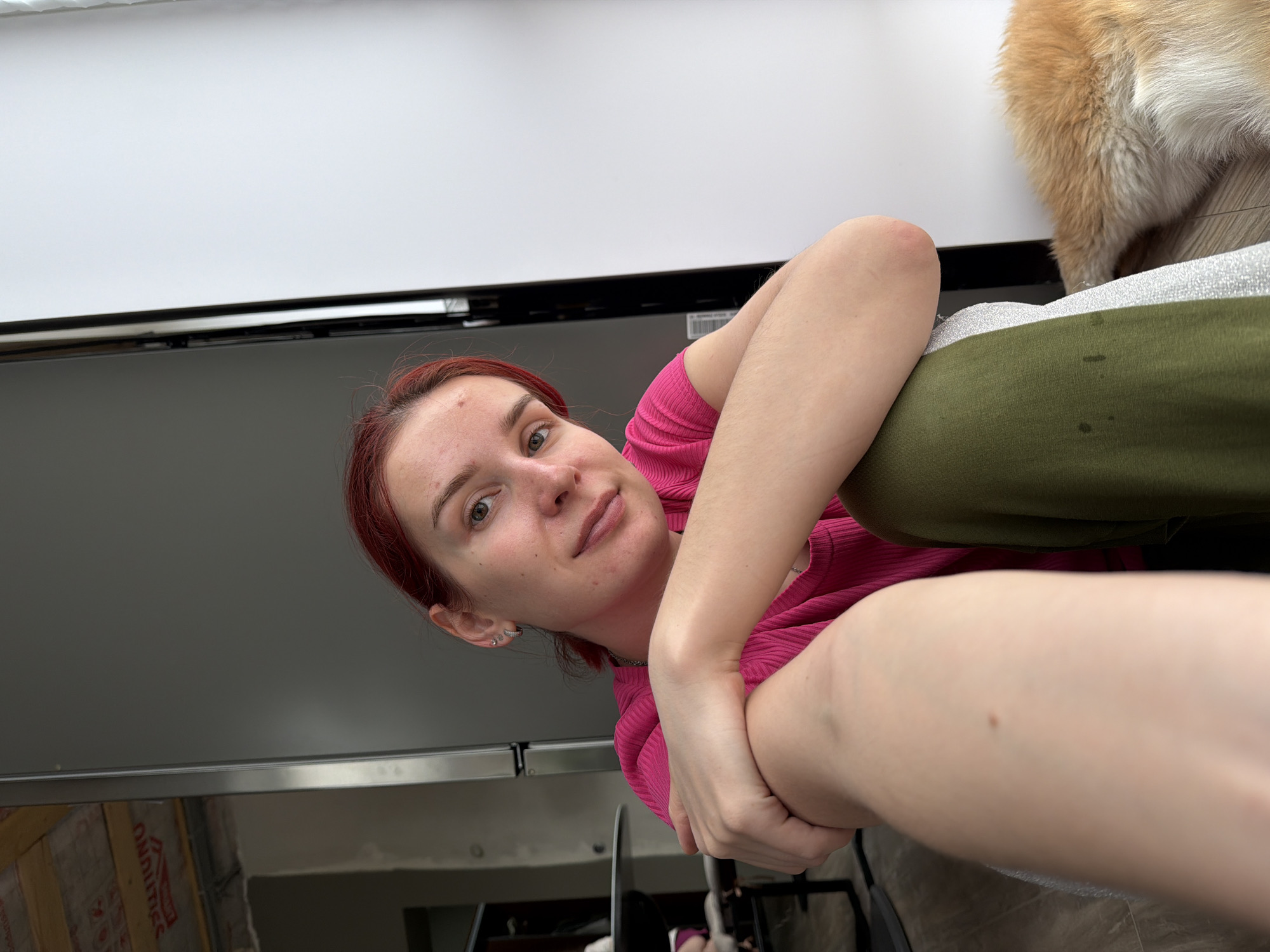A selfie captures a person with red hair and fair skin, wearing a pink ribbed top and green pants. Their arms are crossed in front, and their face is visible. A portion of a dog’s fur is visible in the upper right corner. The background includes a white surface above a black, possibly electronic, object, and the reflection of a cluttered space. The location in Chaikovsky, Russia cannot be determined from the image.