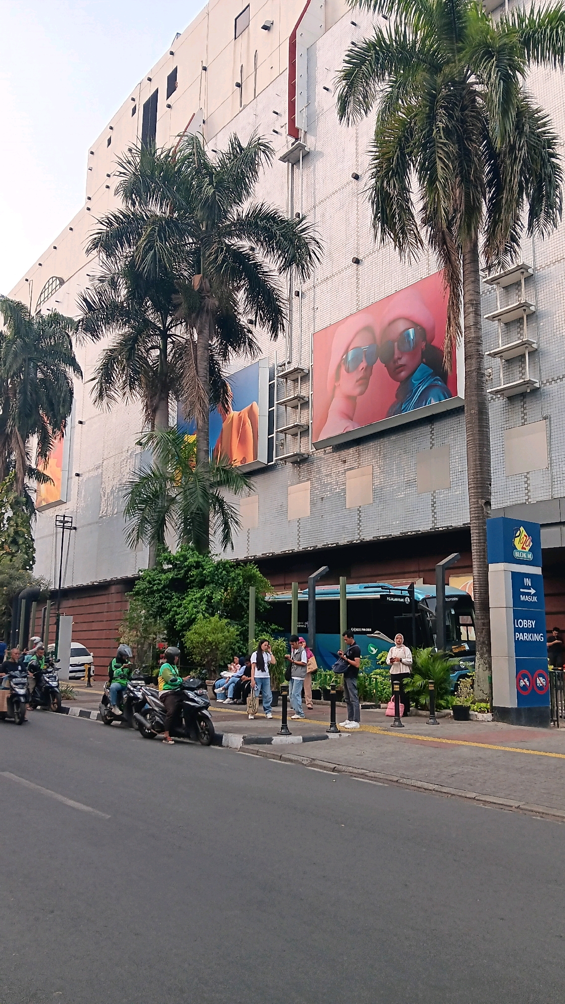 The image depicts the exterior of the Blok M Plaza in South Jakarta, Indonesia. A large building with a tiled facade dominates the background, featuring several large billboards. In the foreground, a street runs alongside a sidewalk where a group of people are standing and sitting. Several motorcycles, some with riders wearing green jackets, are parked along the curb. A blue bus is visible behind a row of potted plants and bollards. A blue and white sign indicates "IN MASUK LOBBY PARKING" with directional arrows and icons. Palm trees are interspersed throughout the scene. The overall atmosphere suggests a busy urban environment.