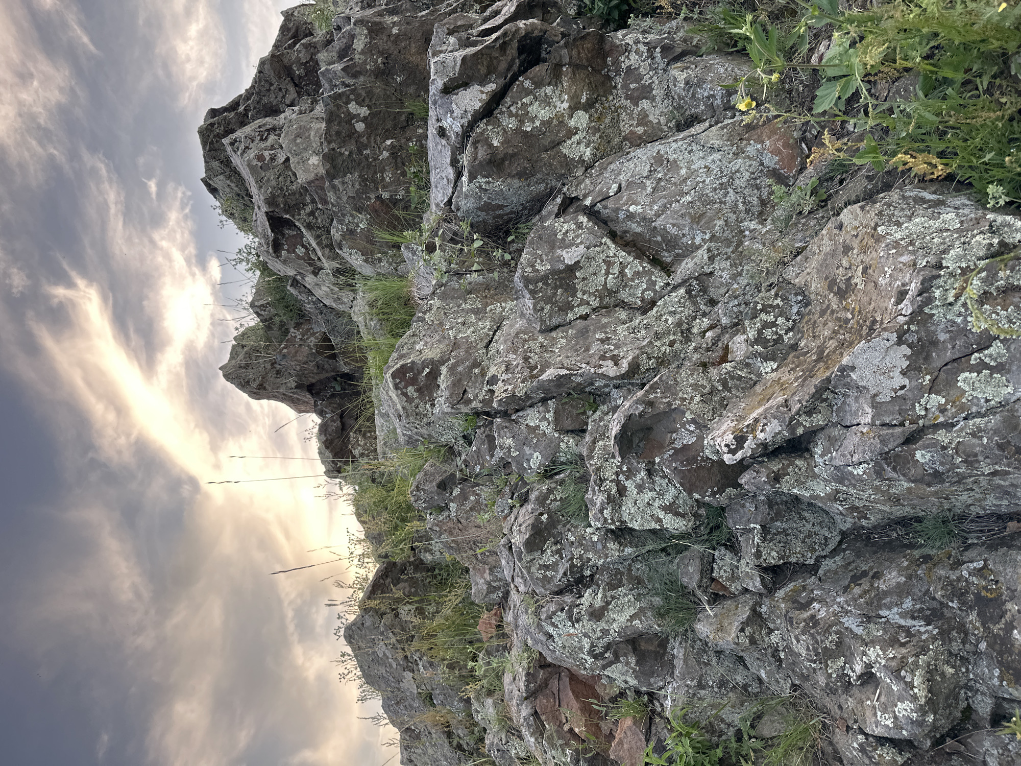 The media file is a close-up, low-angle shot of a rocky outcrop against a cloudy sky. The rocks are dark gray and appear rough and weathered, with patches of pale green lichen. Tufts of dry grass and small green plants grow in crevices between the rocks.
The sky on the left side of the image is dramatic, with streaks of bright light breaking through dark, wispy clouds. The lighting suggests it is either dawn or dusk, with the sun low on the horizon. The overall mood is natural and somewhat rugged. There is no visible text.
