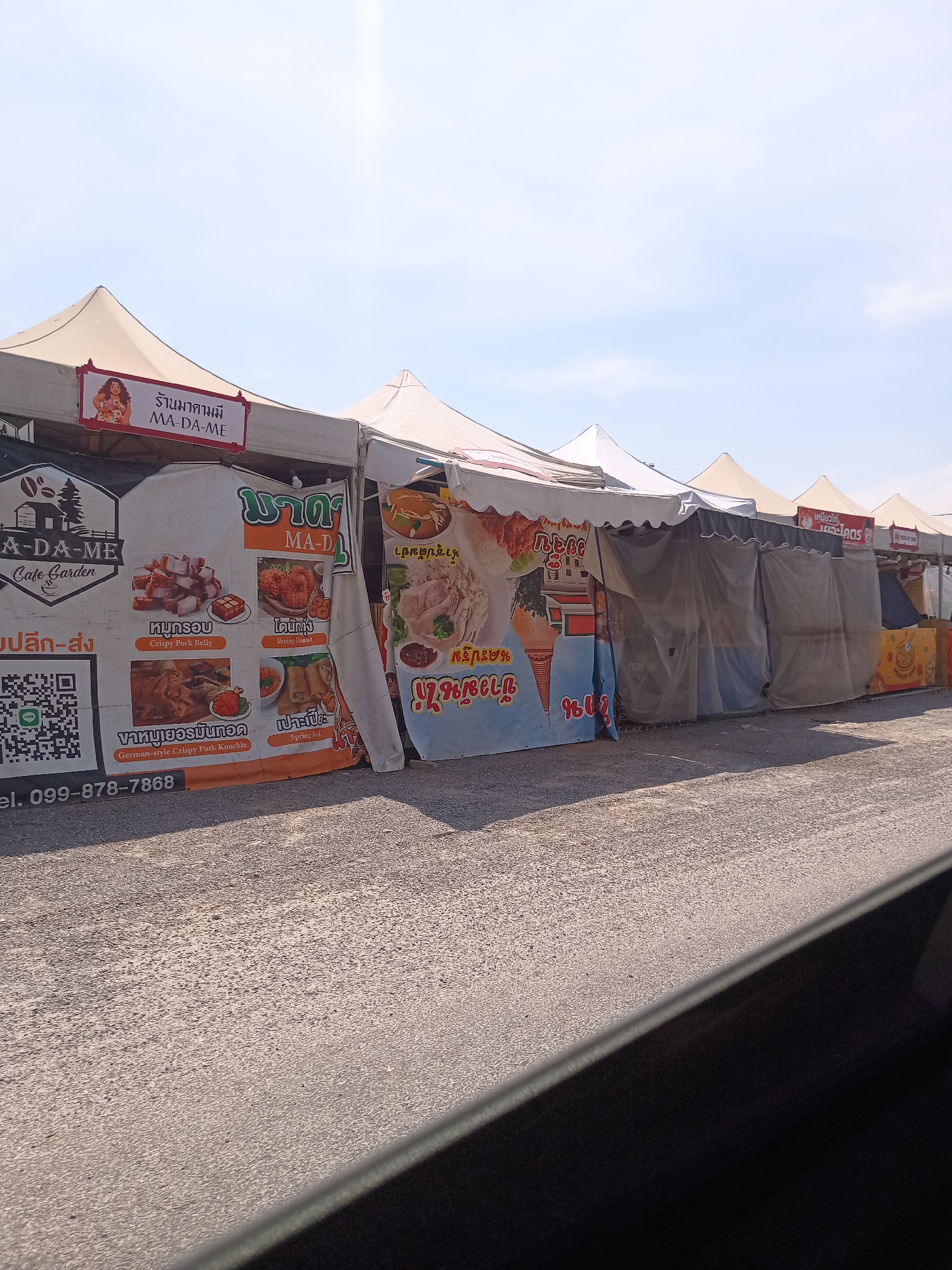 The image captures a row of food stalls under white tent-like structures along a paved road, possibly within Hua Hin City Municipality, Thailand. Each stall is marked with advertising signage displaying various Thai dishes, including crispy pork belly, shrimp donuts, German-style crispy pork knuckle, and spring rolls. The first stall on the left has a prominent sign labeled "A-DA-ME Cafe Garden" with a QR code and a phone number. Another stall features a graphic of an ice cream cone and a text reading “ข้าวมันไก่”. The clear blue sky suggests it's daytime. The image appears to be taken from inside a vehicle, indicated by the car door in the foreground.
