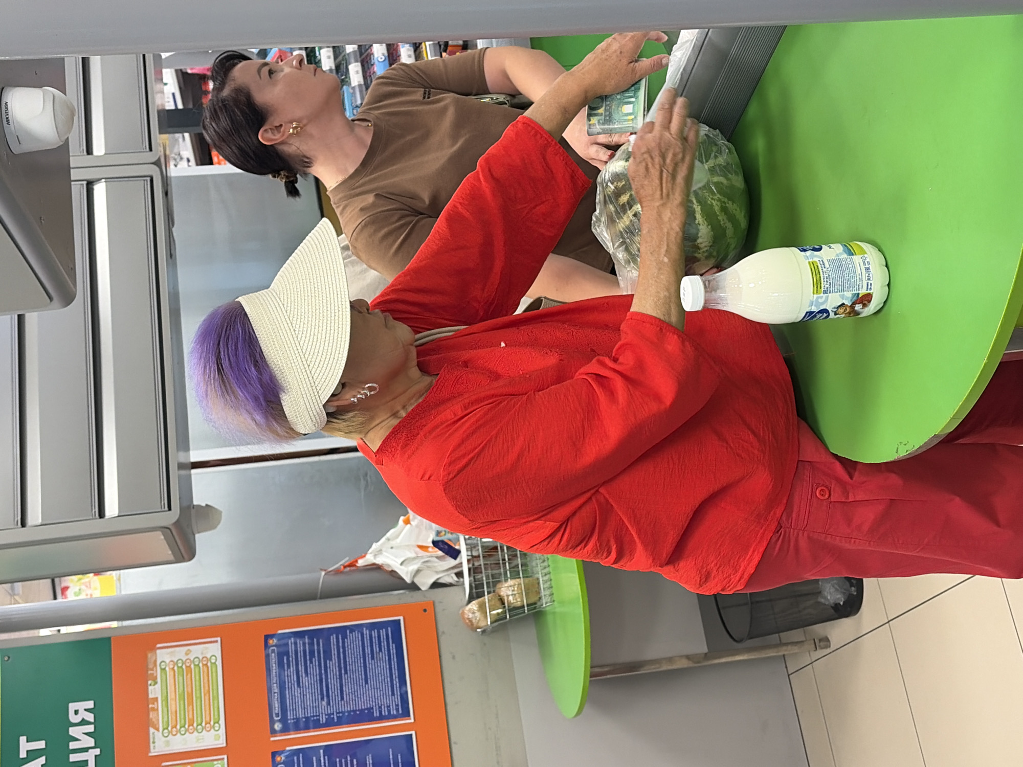 An older woman with vibrant purple hair and a white visor is standing at a checkout counter in a store, paying for her items. She is wearing a bright red shirt and red pants. In front of her, on the green counter, is a bottle of milk. To her right, a smaller woman in a brown shirt is also present. The woman with purple hair is holding a large watermelon wrapped in plastic. The checkout counter appears to be in a grocery store or market, with shelves of products visible in the background.