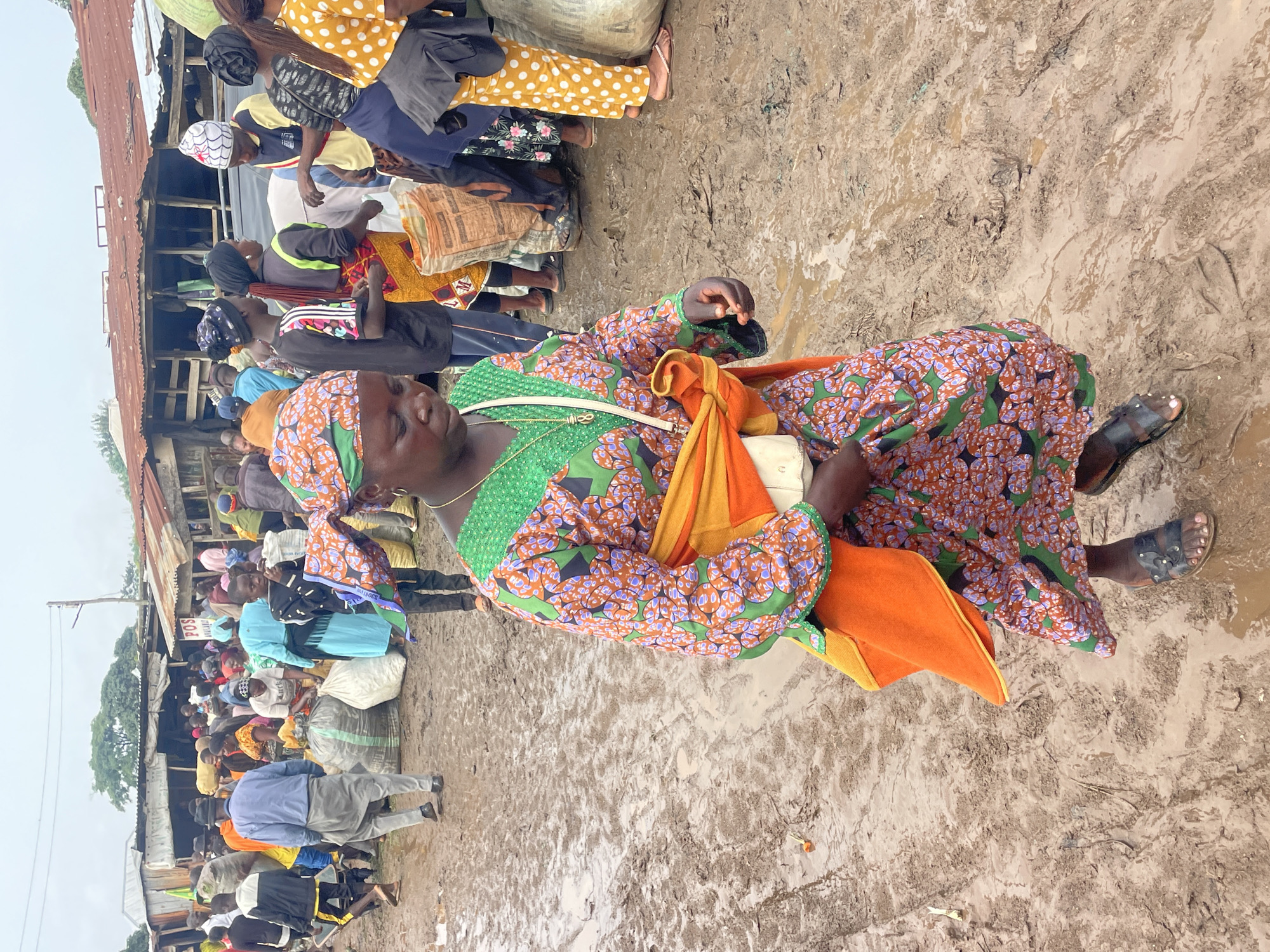 The image depicts a bustling outdoor market in Gura Top, Nigeria, characterized by muddy terrain and crowded conditions under an overcast sky. The foreground is dominated by a woman wearing a vibrant, patterned dress and a green headscarf, with an orange sash tied around her waist. She carries a white bag over her shoulder and is walking through the mud.
In the background, a diverse crowd of people are engaged in various market activities. Many are seen carrying large sacks, possibly containing goods for sale or transport. The market stalls appear rudimentary, constructed from corrugated metal and wood. The ground is heavily saturated with water, indicating recent rainfall or poor drainage. The overall atmosphere suggests a lively and active marketplace despite the challenging environmental conditions.