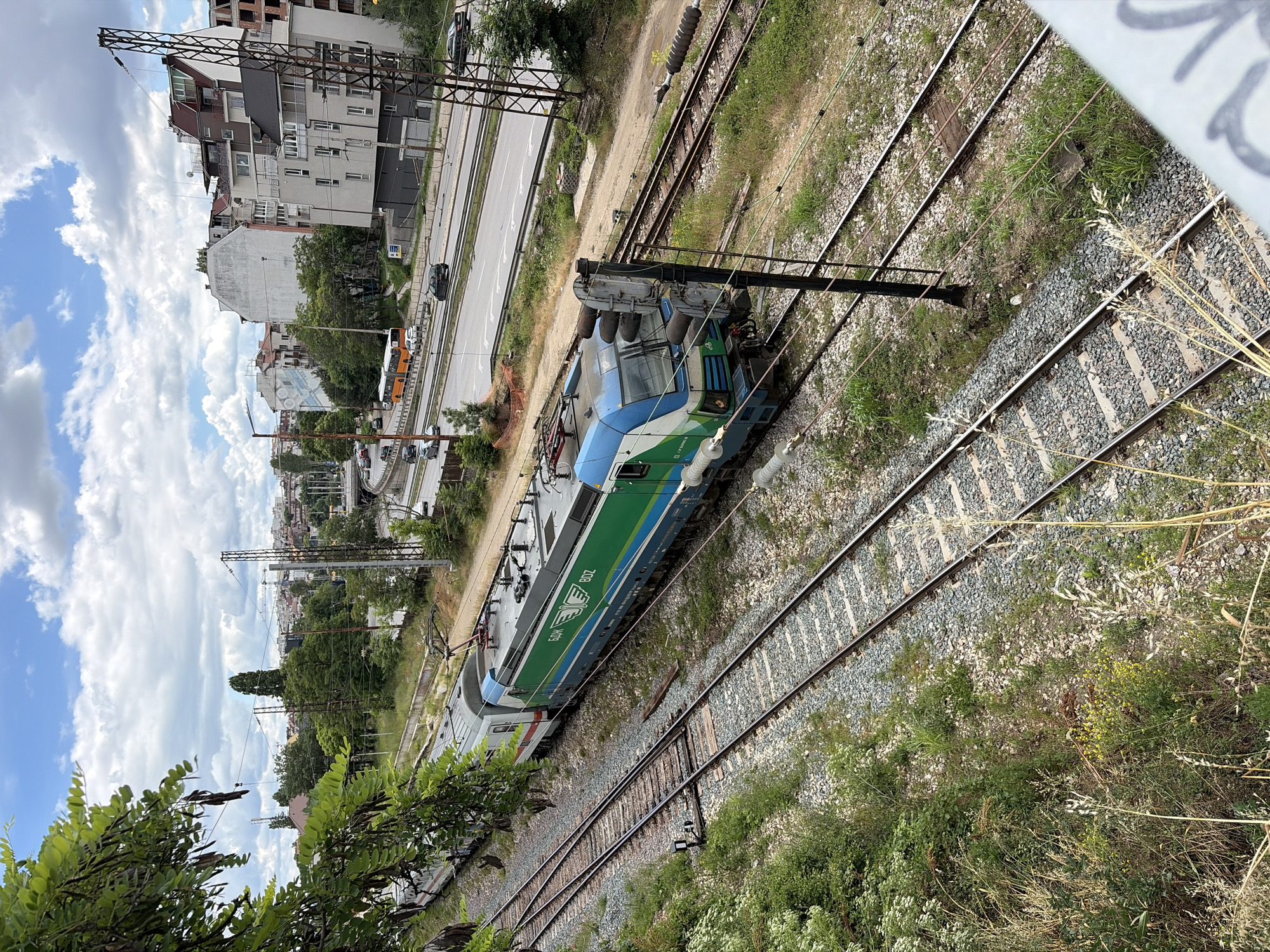 The image shows an electric locomotive, labeled "BDZ", on a railway track in an urban environment, likely in Sofia, Bulgaria. The locomotive is green and blue with white and gold accents. It is pulling several train cars. The railway tracks are surrounded by gravel and overgrown vegetation. In the background, there is a multi-lane road with cars and a bus. Various buildings, including apartment complexes and what appears to be under construction, are visible behind the road. Overhead power lines for the electric train are prominent. The sky is partly cloudy. The scene captures a moment of urban transportation infrastructure in operation, with a train on one set of tracks and vehicular traffic on a nearby road.