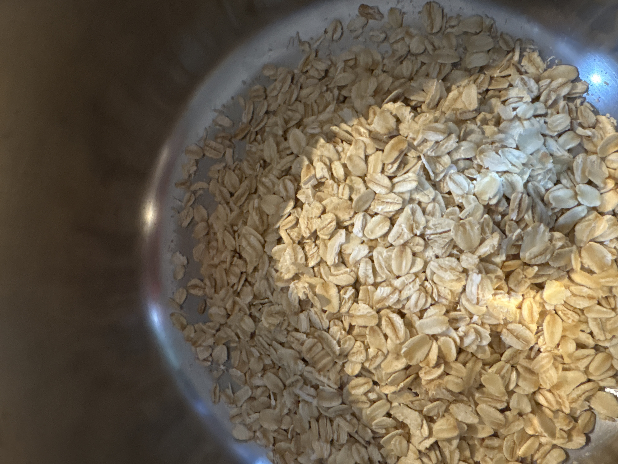 The image shows a close-up view inside a stainless steel bowl filled with rolled oats. The oats are densely packed and appear dry. The lighting is uneven, with some areas brighter than others. The bowl takes up most of the frame. There is no indication of the location being Karon, Thailand. There are no people, actions, or interactions visible.