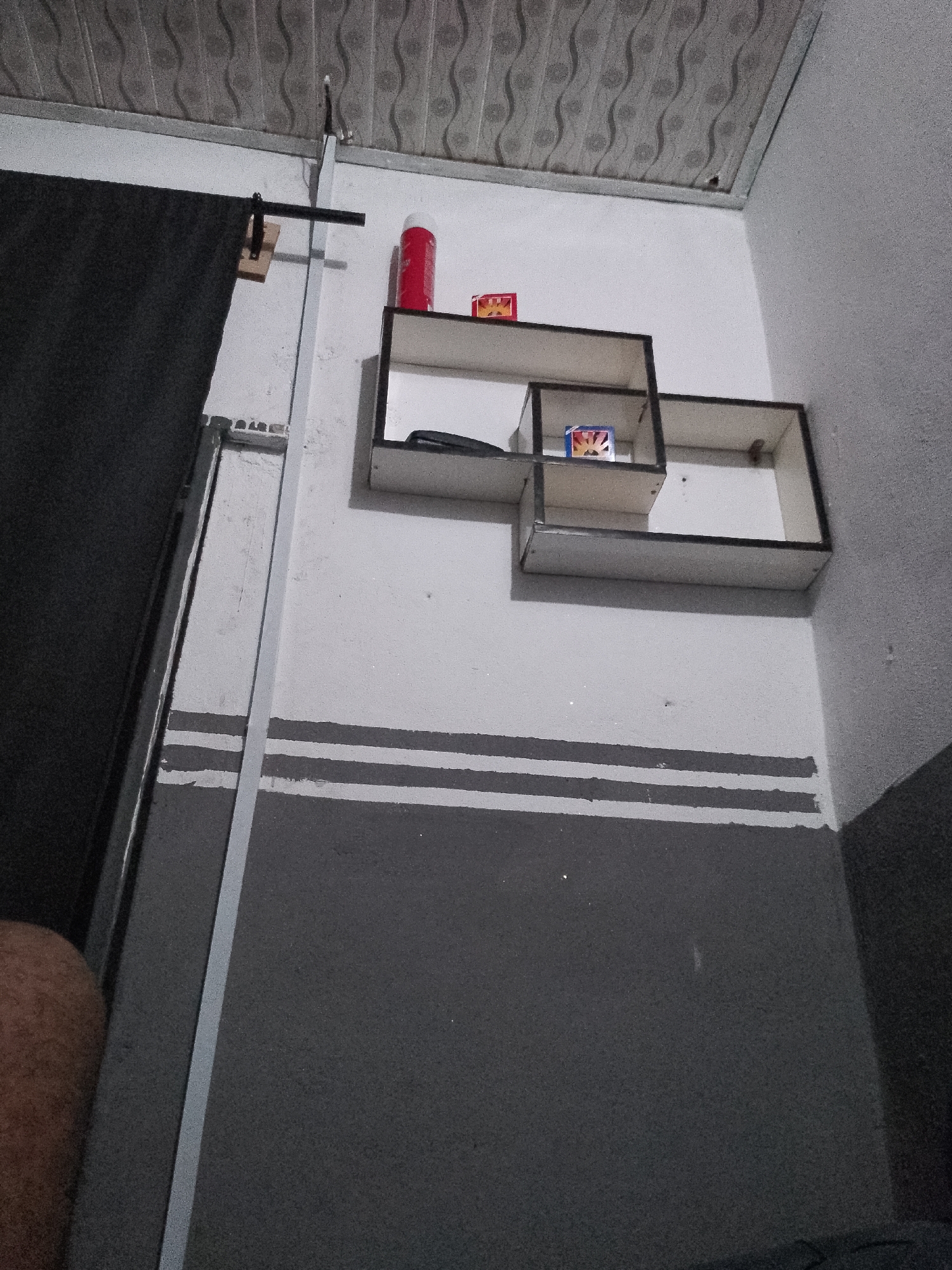 In Jauro Yinu, Nigeria, an indoor scene depicts a wall with a ceiling that has a patterned texture. A metal shelf unit is mounted on the wall, featuring three cubbyholes. A red cylindrical object, possibly a can, is positioned on the top shelf, alongside two small rectangular boxes. A black object, appearing to be a phone, rests on the middle shelf. The wall is painted with a horizontal stripe pattern in shades of grey. To the left of the wall is a dark curtain and a tall, thin vertical metal support. A person's thigh is visible in the lower left corner of the frame.