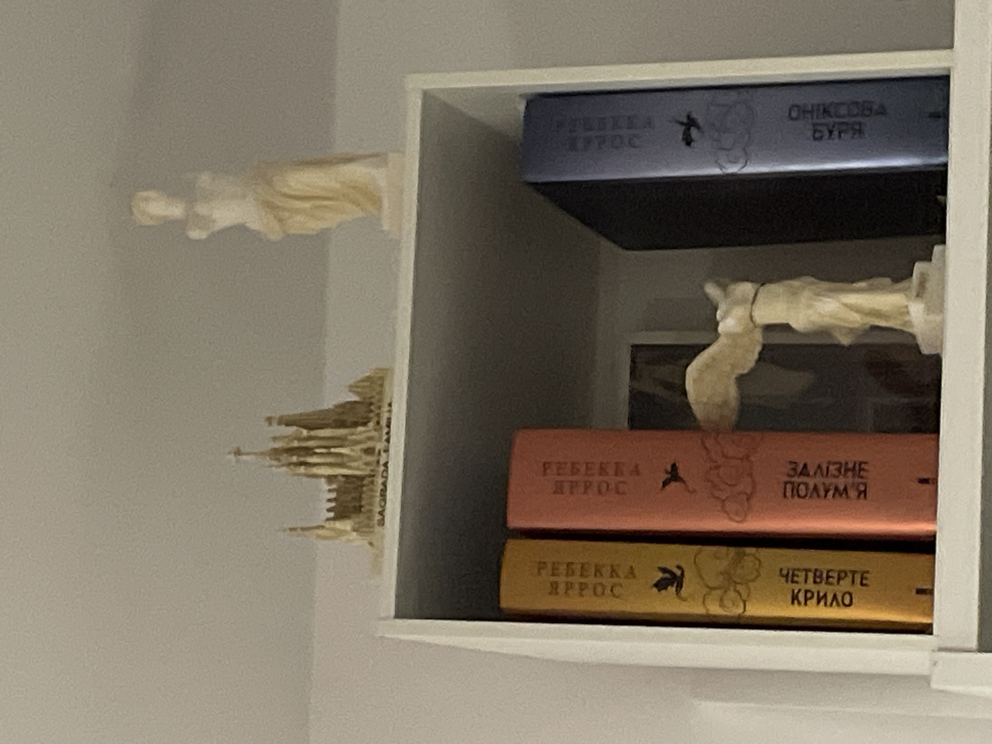 The image shows a white bookshelf with several objects on it. On the top shelf, there is a miniature model of a cathedral on the left and a statue of a female figure on the right. The cathedral model has "SAGRADA FAMILIA" written on its base. The female figure appears to be draped in cloth.
On the lower shelf, there are three books. The book on the far left is yellow with black text and an illustration of a bird and swirling patterns. The second book from the left is orange-pink with black text and similar illustrations. The third book from the left is dark blue with light blue text and an illustration of a bird and swirling patterns. Next to the books on the lower shelf is a small statue of a winged female figure, possibly Nike of Samothrace. Behind the books and the winged statue is a framed picture or artwork, mostly obscured by the books. The background of the image is a plain, light-colored wall. The lighting is soft and appears to be from an overhead source.