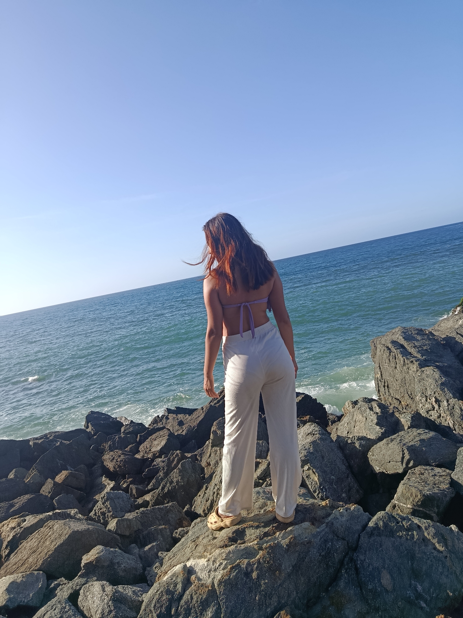 A woman with long, reddish-brown hair stands on a rocky coastline, facing away from the camera and towards the ocean. She is wearing a light purple bikini top and loose-fitting, off-white pants. The ocean is a deep blue with white-capped waves crashing against the rocks. The sky is clear and a pale blue. The rocky terrain is composed of large, dark gray boulders of various sizes. The image appears to have been taken on a sunny day. This scene is located in the city of Pedro González, Venezuela, known for its beautiful coastal landscapes.