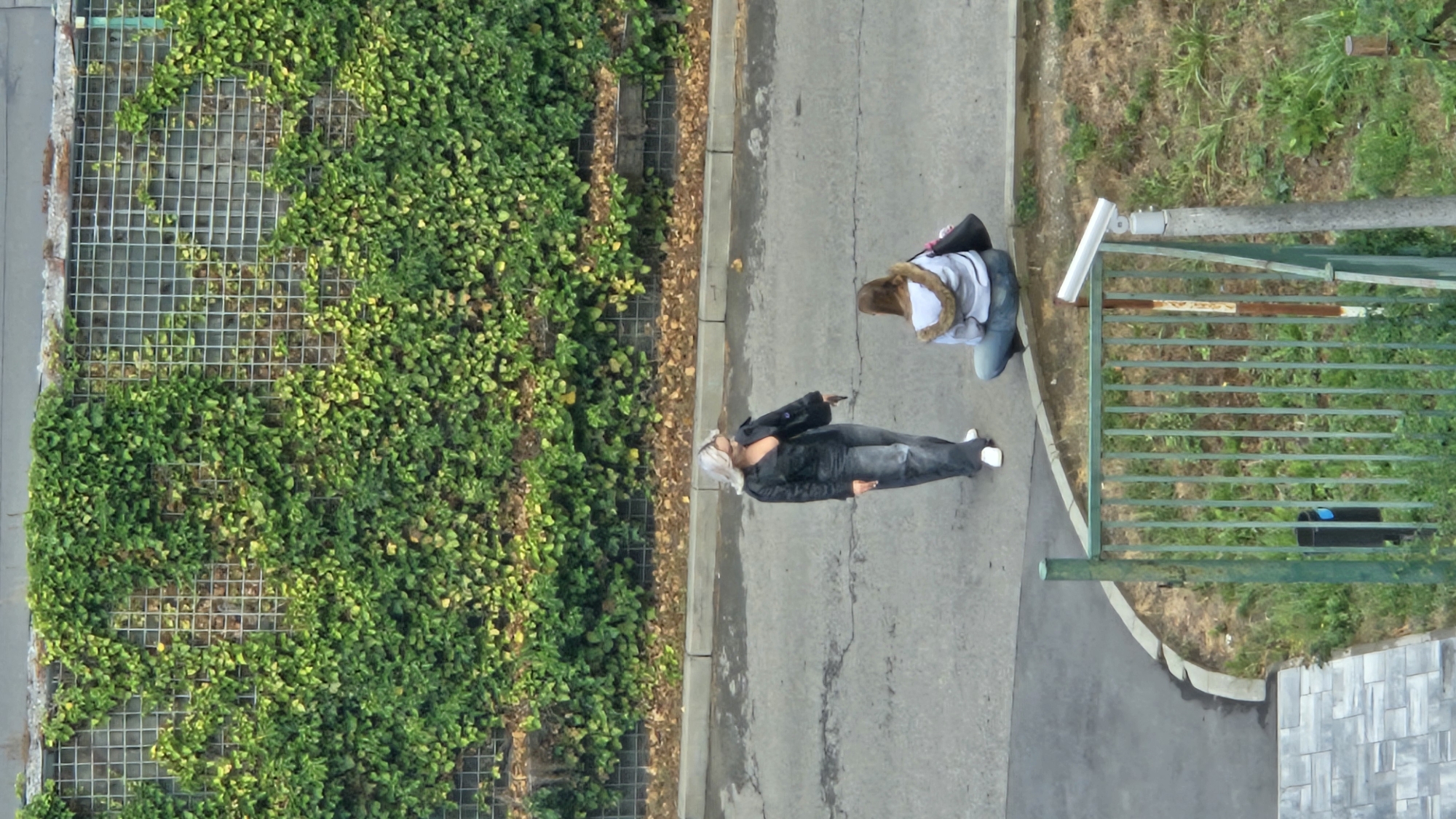 Two women are standing and sitting on a sidewalk near a chain-link fence covered in green ivy. The woman on the left is standing and wearing a black leather jacket, dark pants, and white sneakers. She has blonde hair and is holding a black purse and a phone. The woman on the right is sitting down, wearing a light-colored hooded jacket with a fur trim and blue jeans. She has brown hair and is carrying a black purse. In the background, a wall is covered with ivy, and a street is visible in front of the sidewalk. A security camera is mounted on a pole near the fence in the foreground. The location is identified as Prešov, Slovakia.