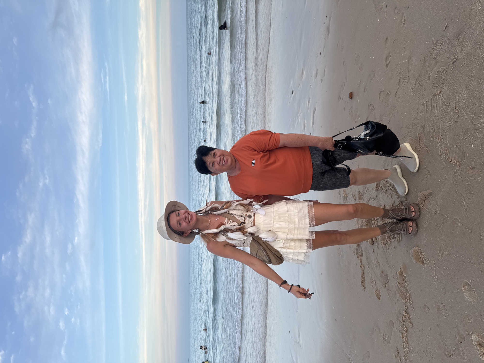 Two individuals are standing on a sandy beach in Phu Thuy Ward, Vietnam, with the ocean and sky in the background. The person on the left is a slender woman with blonde hair, wearing a white bohemian-style dress, a wide-brimmed hat, and large feathered earrings. She is also wearing brown sandals and a crossbody bag. The person on the right appears to be an older Asian woman with short dark hair, wearing an orange t-shirt and dark shorts, with grey sneakers and carrying a black bag. Several people are visible in the water in the distance, and the sky displays a gradient of blue and pink hues, suggesting it is either early morning or late evening. Footprints are visible in the sand around the individuals.