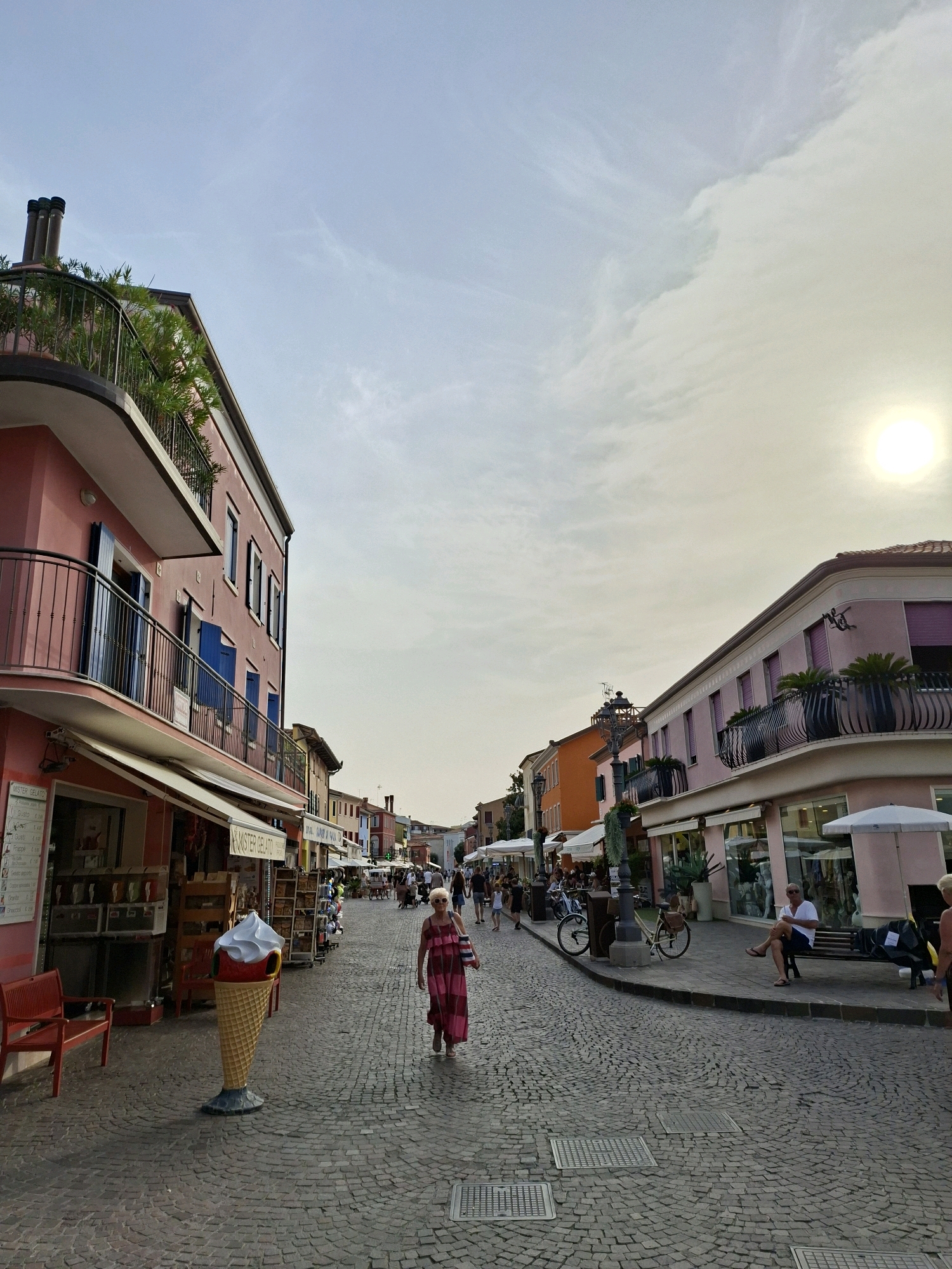 The image depicts a cobblestone street in Caorle, Italy, lined with colorful buildings. The architecture features pastel hues, balconies with wrought-iron railings, and awnings extending over shops. A large, decorative ice cream cone sculpture stands prominently on the left side of the street, near an establishment labeled "MISTER GELATO."
Several people are visible, engaged in various activities. A woman in a pink and red patterned dress is walking down the center of the street. On the right, a man in a white t-shirt and dark shorts is seated on a bench outside a shop. Other individuals are seen walking, browsing in shops, and sitting at tables in the distance.
The street is paved with interlocked stones and has some metal grates on the ground. The sky is partly cloudy with a visible sun, suggesting it might be late afternoon or early evening. The overall atmosphere appears to be that of a pleasant, lively tourist area.