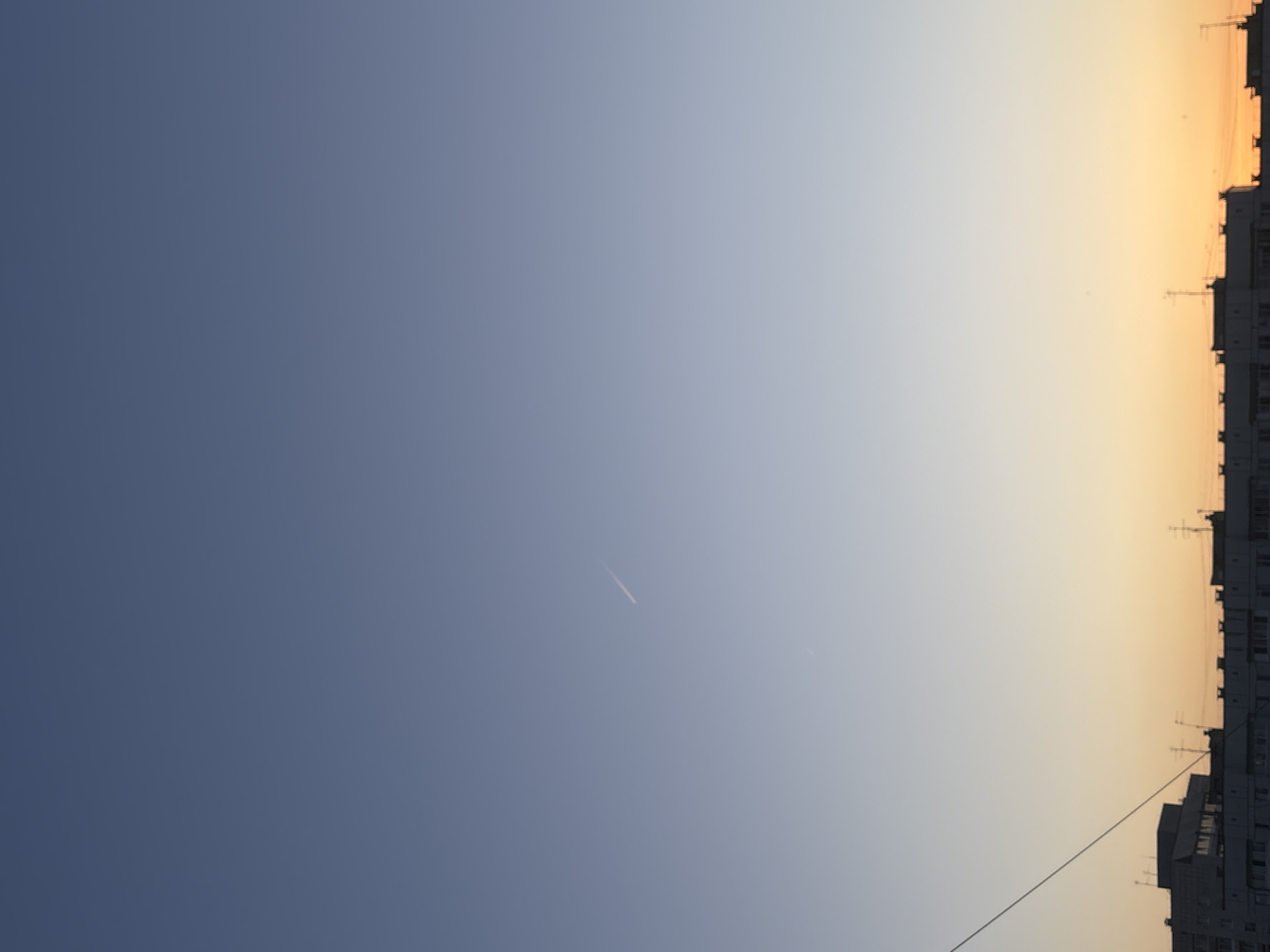 The image depicts a view of the sky over a city, presumably Chaikovsky, Russia. The sky transitions from a deep blue on the left to a lighter, more yellowish hue on the right, likely indicating either sunrise or sunset. Silhouetted against the sky on the right side are the outlines of apartment buildings, complete with various antennas and power lines. A faint contrail is visible in the upper-center portion of the image.