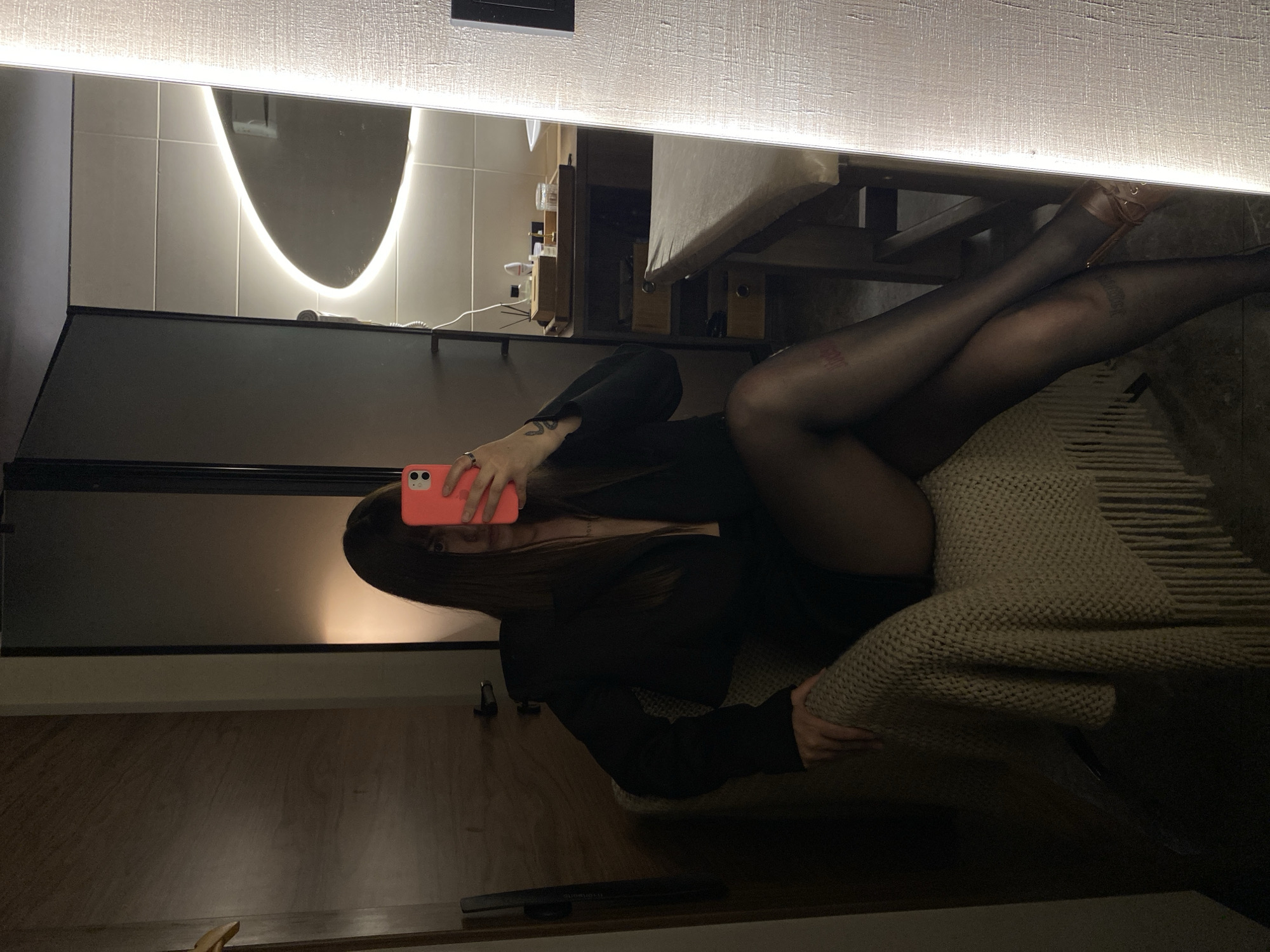 A woman in black pantyhose, high heels, and a black blazer is reclining in what appears to be a hotel room or apartment. She is holding a pink phone in front of her face, likely taking a selfie in a mirror. The room has modern decor. The location is unknown, as there are no indications of Votkinsk, Russia.