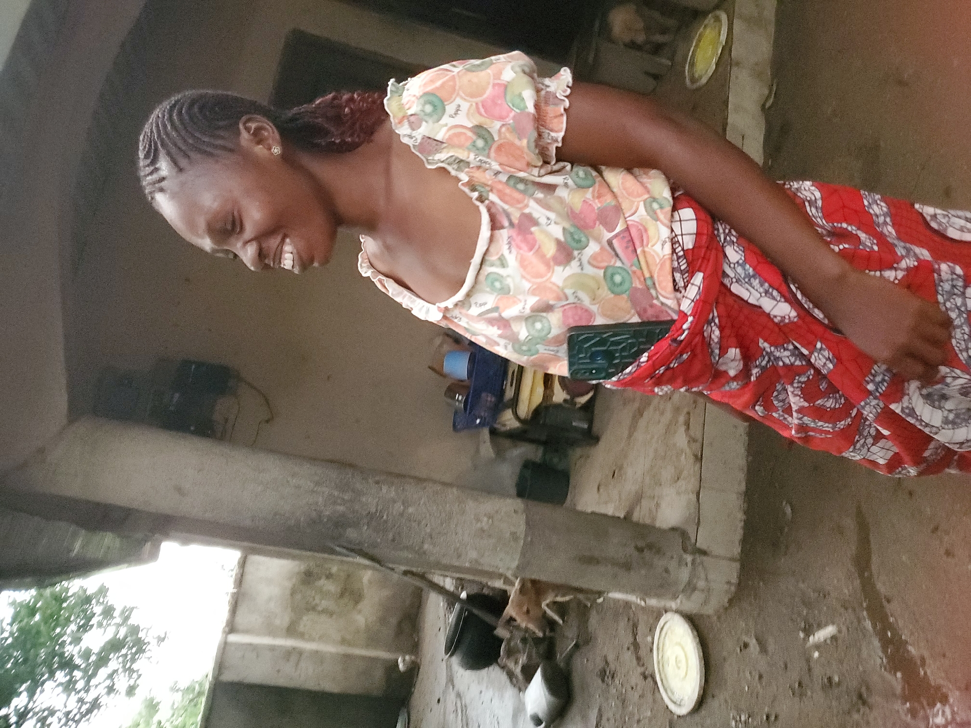 In Mararaba, Nigeria, an adult Black woman with braided hair is smiling and looking down. She is wearing a colorful top with fruit patterns and a red-patterned fabric wrapped around her waist. She is standing near a concrete structure. In the background are various objects including containers and other household items. The ground is wet.