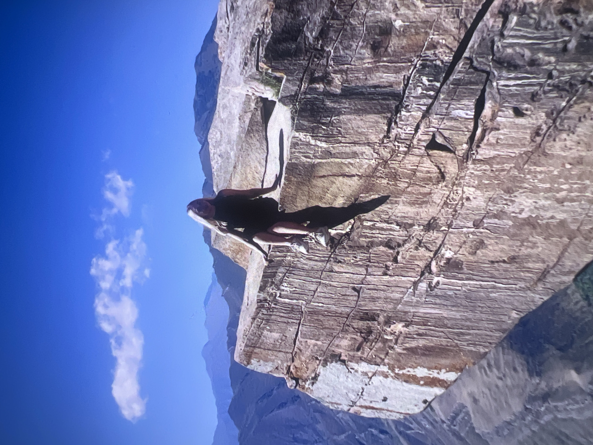 The media file captures a high-angle view of a person lying on a vast, rugged rock ledge, set against a bright blue sky and distant mountain ranges. The image is initially rotated 90 degrees clockwise.
**Subjects:** A single person, likely an adult, is the primary subject. They are wearing dark clothing, appearing to be a dark top and shorts or leggings, and are lying on their back on a flat section of the rock. Their head is visible, and their body casts a long, distinct shadow across the rock surface. The main object is the massive, layered rock formation that constitutes the cliff and ledge. The scene encompasses a majestic mountain landscape.
**Activity or event taking place:** The person appears to be resting, relaxing, or posing for a photograph on the precarious rock ledge. The context suggests an outdoor activity such as hiking, mountaineering, or exploration at high altitude.
**Notable details:** The time of day is clearly bright daylight, with strong, direct sunlight creating sharp shadows. The sky is a clear, vibrant blue with a few scattered, wispy white clouds. The rock formation exhibits clear horizontal stratification and vertical fissures, indicating a natural, geological setting. Distant mountains, rendered in muted blue tones, suggest a vast and expansive environment, implying a high-altitude location. No specific emotions are discernible on the person's face due to the distance and angle.
**Visible text:** There is no visible text in the image.