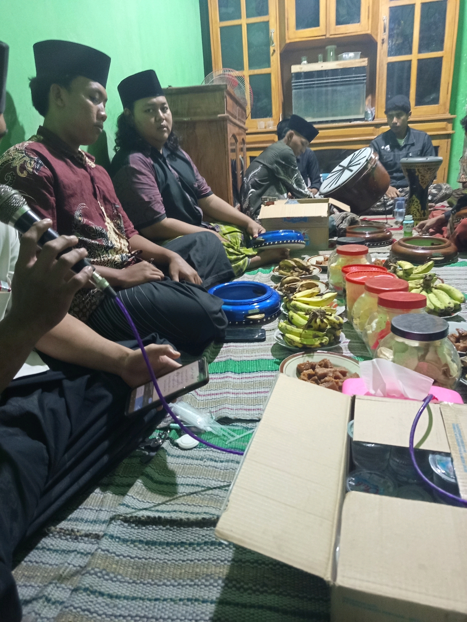 The image depicts a gathering of people in what appears to be an indoor setting, possibly a home in Mrican, Indonesia. The atmosphere suggests a casual or informal event, with several individuals seated on the floor on a woven mat. The lighting is dim, indicating it might be evening or nighttime.
The foreground focuses on a person holding a microphone and a smartphone, suggesting they might be leading a song, prayer, or a presentation. To their left, another person is also seated, wearing a maroon patterned shirt and a black peci (a traditional cap). Another individual to the right is wearing a dark shirt and also a peci. In the background, more people are visible, some engaged with musical instruments, including what looks like a frame drum and a goblet drum.
The scene is set around a spread of food and drinks. Plates of bananas are prominently displayed, along with several jars containing food items, and a bowl of what seems to be cubed snacks. A cardboard box is also in view, containing more items. Blue and red containers, possibly for food or drinks, are also present. The overall impression is of a communal gathering, possibly a religious or cultural event, where food and music are shared. The people are dressed in modest attire, fitting for the context of a gathering in Indonesia. The background shows wooden furniture, possibly cabinets, suggesting a domestic environment. There is no visible text in the image.