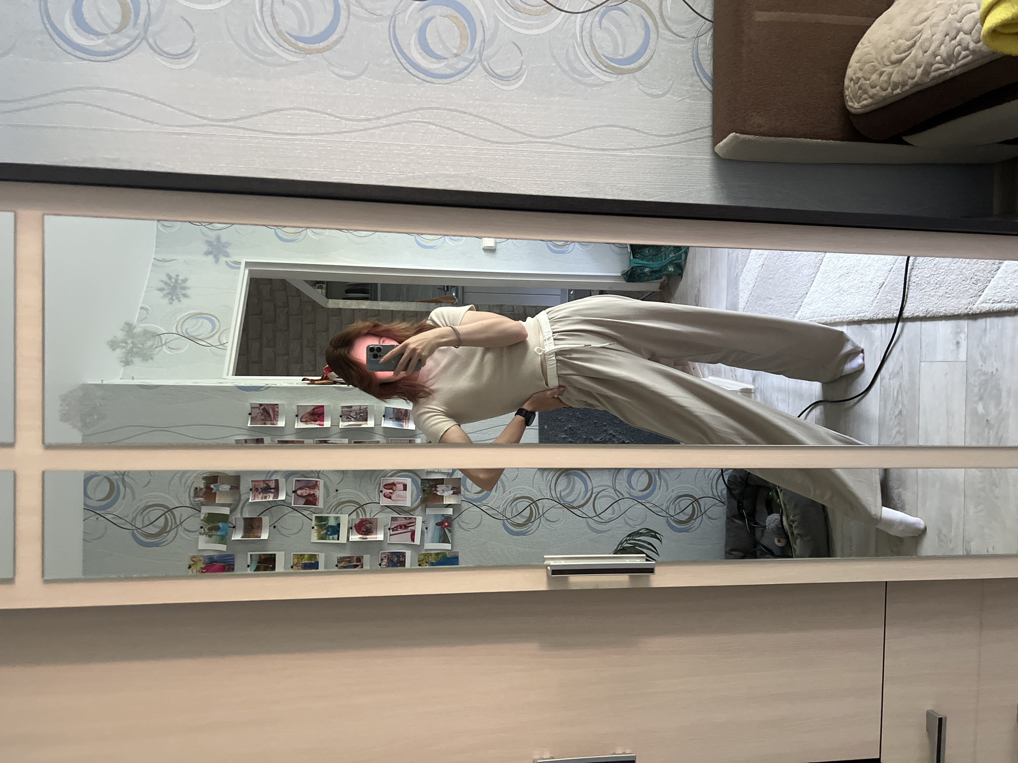 The image shows a woman standing in front of a mirror, taking a selfie. She is wearing a beige t-shirt and beige wide-leg pants. The mirror reflects her and the room behind her. The room appears to be a bedroom, with a bed visible in the background. There are also several framed photos on the wall and a window. The woman is holding a smartphone in her right hand and appears to be posing. The overall scene suggests a casual, personal moment captured in a domestic setting. The location is identified as Chaikovsky, Russia.