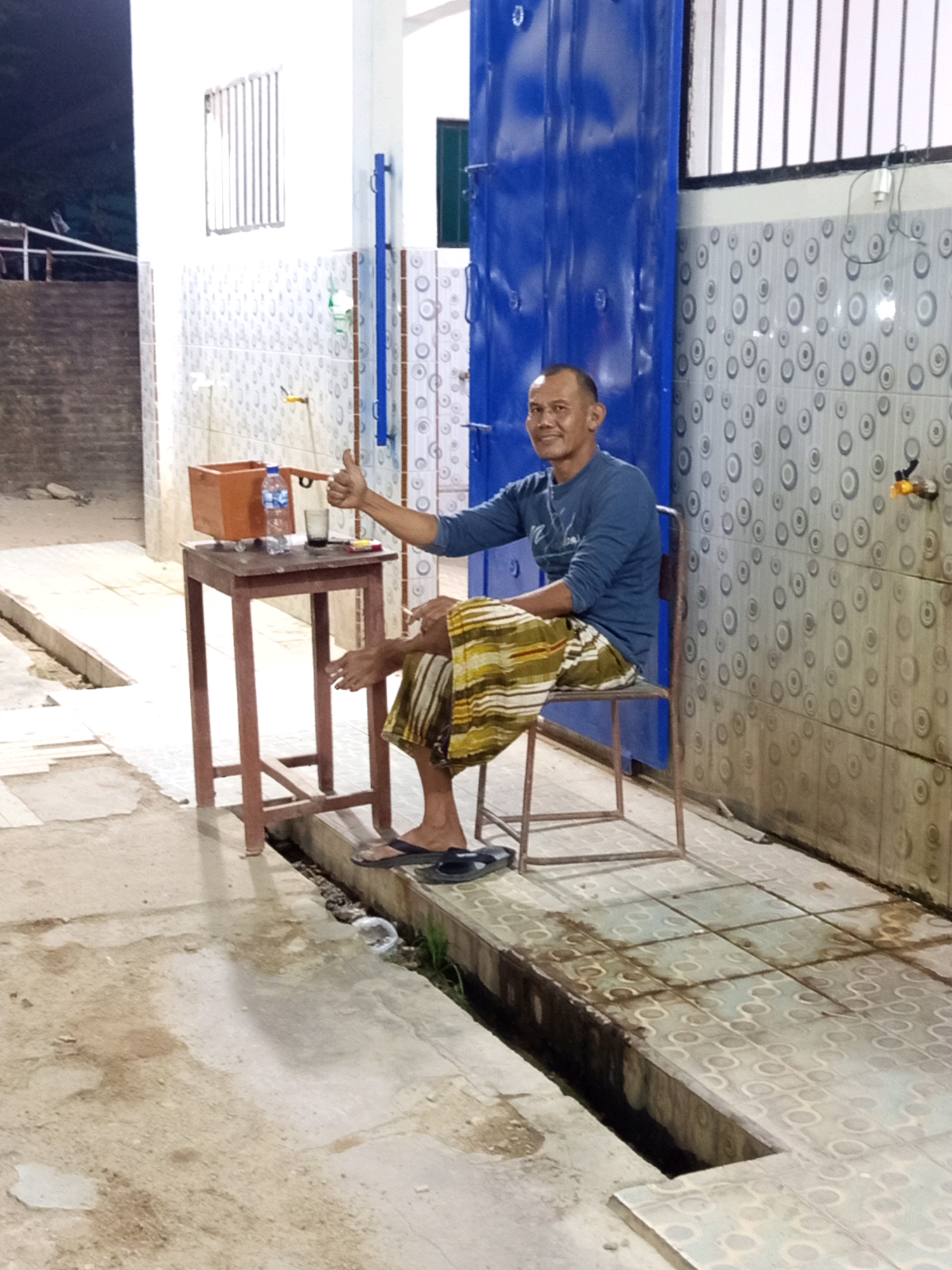 The image shows an adult male sitting on a chair next to a small table with a drink and a bottle of water. The man is wearing a long-sleeved blue shirt and a sarong. He is holding a cigarette in his right hand and giving a thumbs-up with his left hand. The table is made of wood and has a rectangular orange container on it. In the background, there is a blue metal door and a wall with patterned tiles. The ground is concrete, and there is a drainage ditch running along the side. The scene appears to be outdoors, possibly in a public area. The overall lighting suggests it might be late afternoon or evening. The image is taken in the city of Pulung Kencana, Indonesia.