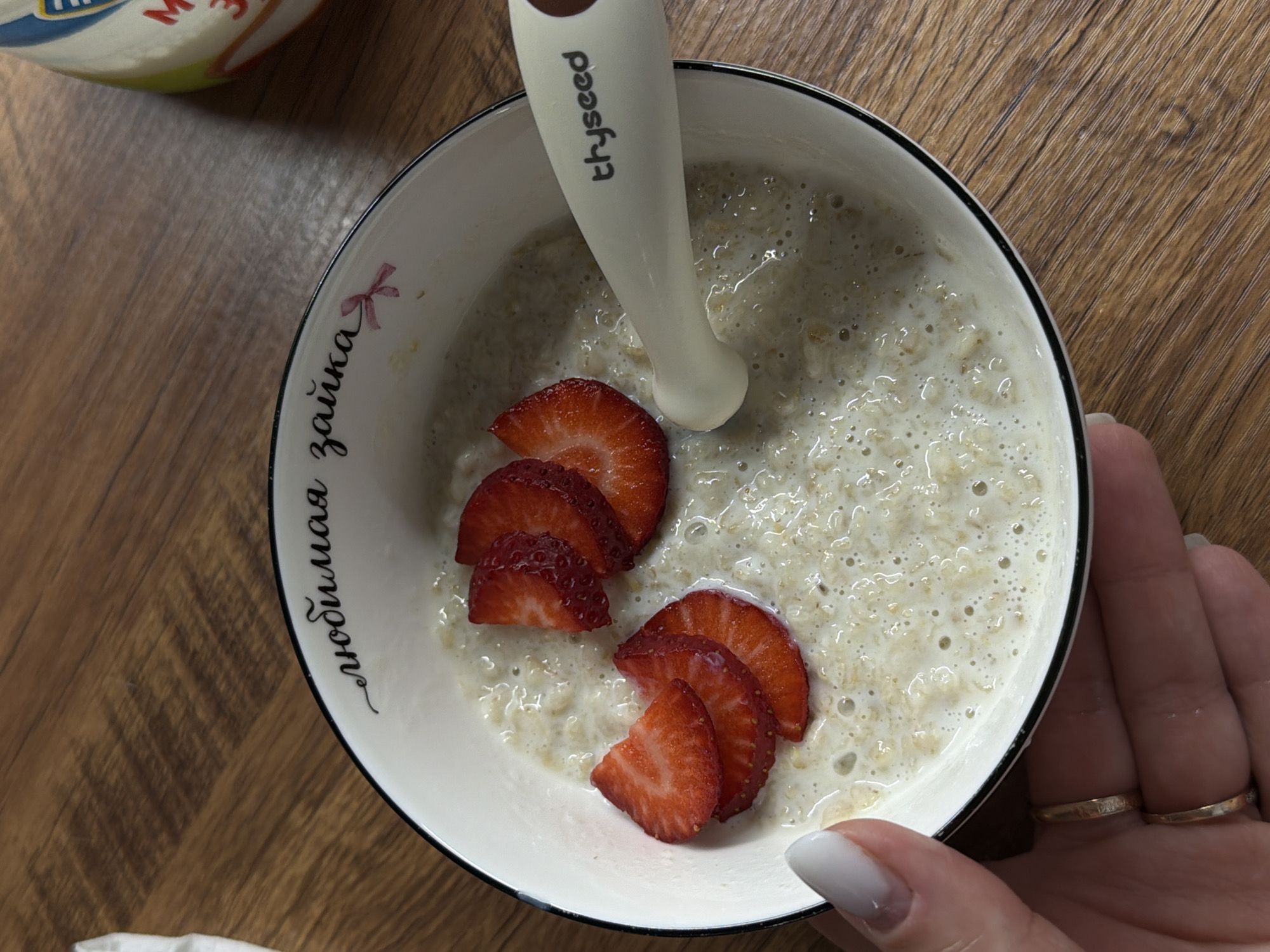A high-angle, close-up shot features a bowl of oatmeal with sliced strawberries on a wooden surface. A white spoon with the word "thyseed" imprinted on it rests in the oatmeal. The bowl is white with a black rim, and the phrase "любимая зайка" (darling bunny) is written in cursive along the side with a small pink bow. A hand with white manicured nails and two gold rings is partially visible, holding the bowl. Another object, possibly a container, is partially visible in the upper left corner of the frame. The scene suggests a breakfast or snack setting, possibly in a home environment. No information indicates the location is Reshetnikovo, Russia.
