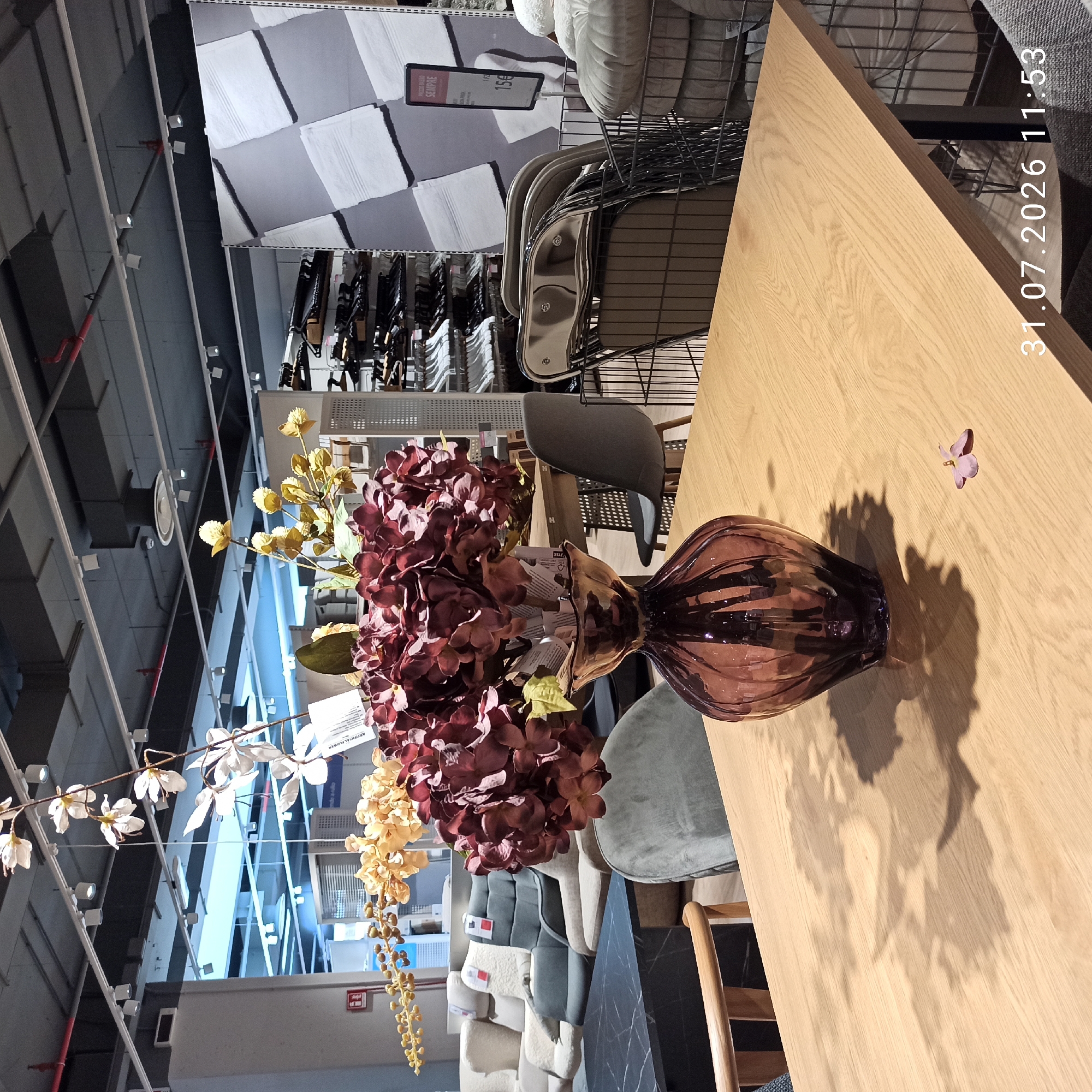 An indoor shot depicts a furniture store with various items displayed. In the foreground, a wooden table holds a dark purple glass vase with artificial flowers, including burgundy hydrangeas and yellow-gold berries, and a single fallen pink flower petal. The background reveals a retail environment with upholstered seating, including chairs and sofas, on display. Further back, clothing racks with hangers are visible, as are sections of shelving with what appear to be towels or linens. The store's ceiling features a grid of lights and industrial elements. The scene is illuminated by artificial lighting. A date and time stamp "31.07.2026 11:53" is superimposed on the lower right corner. The location is identified as Modugno, Italy.