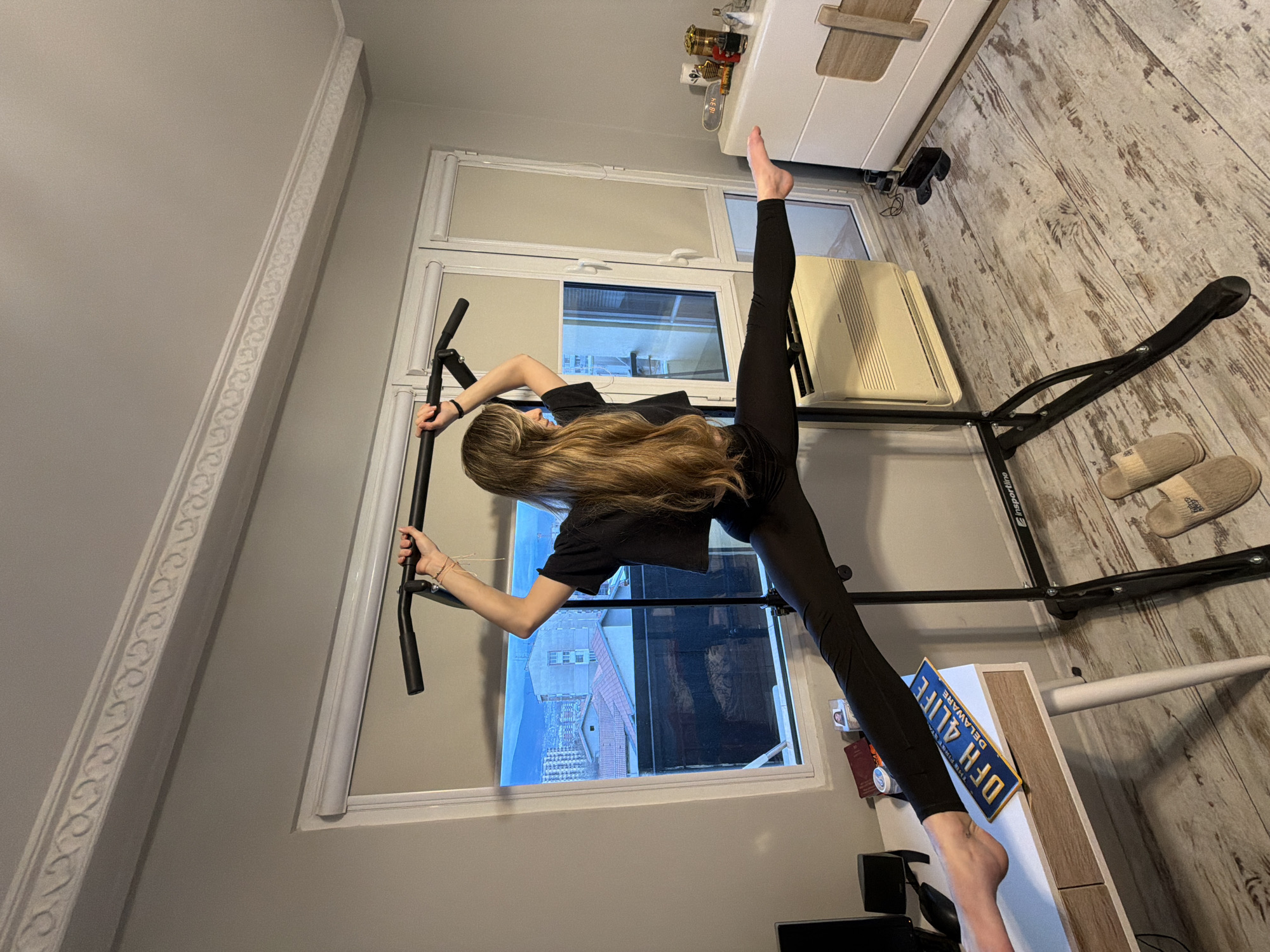 A young woman with long blonde hair is performing a split inside a room. Her right leg is extended vertically upwards towards the top of the frame, while her left leg extends horizontally towards the left side. She is holding onto a black pull-up bar attached to the window frame.
The room has white walls and wood-patterned flooring. A window with a view of buildings is in the background. A small television or monitor is present towards the bottom of the frame. To the right, there is a piece of exercise equipment and a cabinet. A blue and yellow Delaware license plate with "DFH 4 LIFE" printed on it is placed on top of the cabinet. A pair of slippers is also present on the floor near the exercise equipment.
Based on the architectural style and the visible cityscape, this image is likely not in Plovdiv, Bulgaria. The buildings appear more modern than what is typically found in the older parts of Plovdiv, and there are no immediately identifiable landmarks.