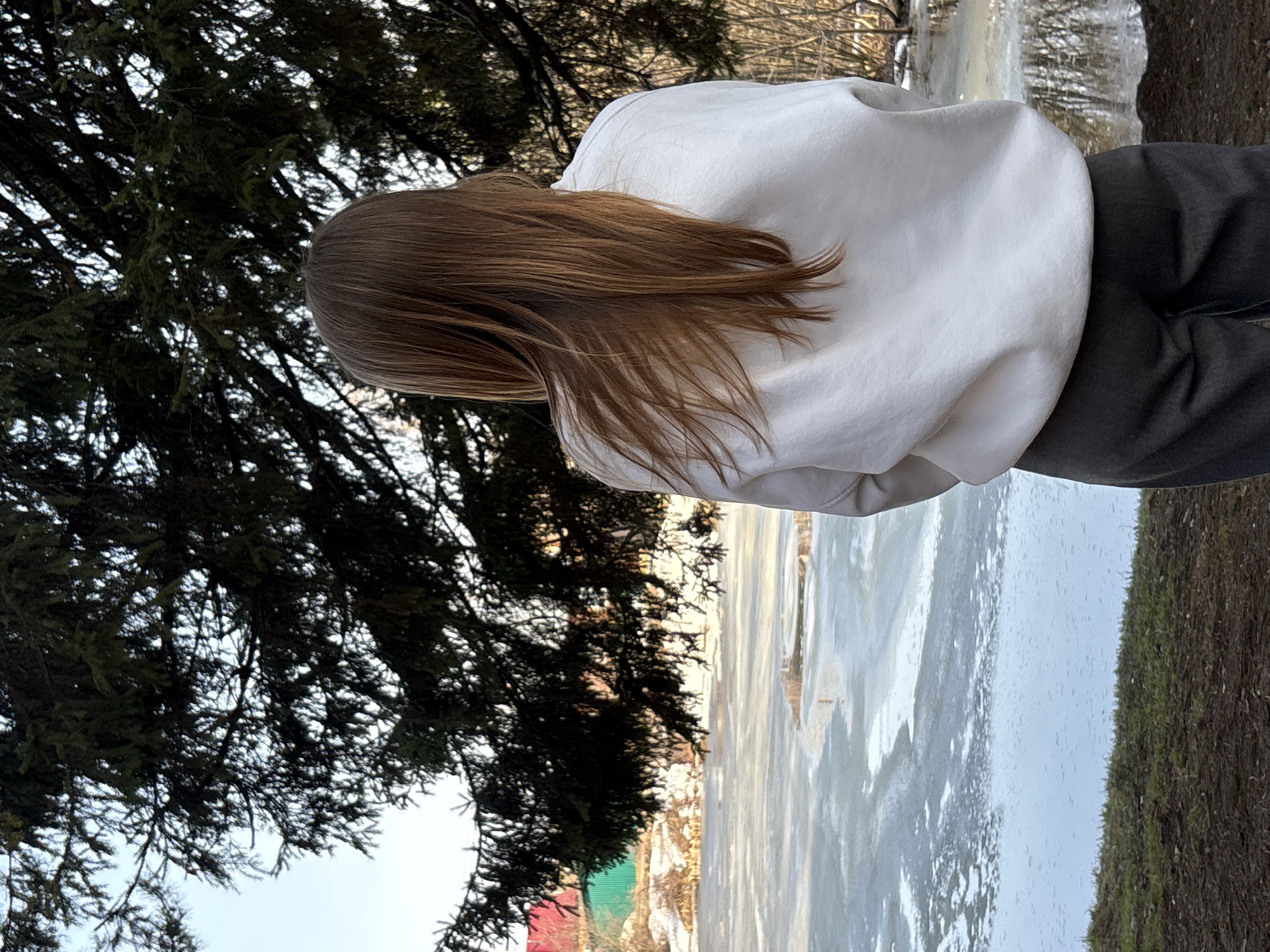 A person stands near an icy surface, possibly a frozen body of water, in Chaikovsky, Russia. They are wearing a white sweatshirt and dark pants. The person is seen from behind, with long brown hair. A dark green tree looms to the left of the frame. The area is snowy and icy, suggesting winter. The ground is covered in a mix of snow, ice, and brown vegetation.