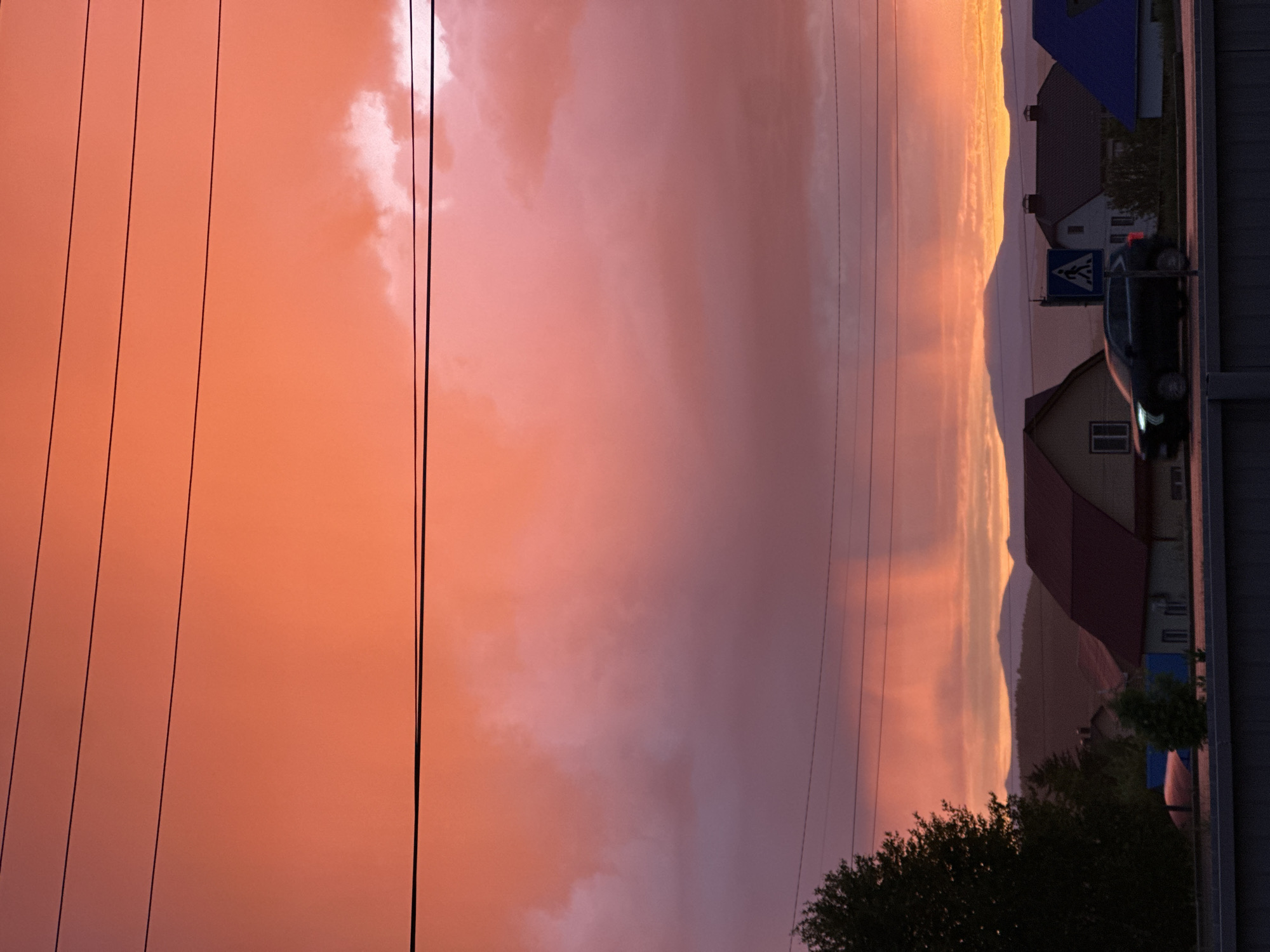 This image captures a vibrant sunset or sunrise over a suburban or rural landscape, showcasing a dramatic sky and distant mountains. The sky is a dominant feature, filled with an intense palette of colors ranging from bright orange and yellow on the left, where the sun appears to be, transitioning into soft pinks and purples across the expanse of clouds. Sunlight breaks through the clouds in various spots.
Several dark power lines stretch horizontally across the sky, adding an element of man-made infrastructure to the natural spectacle. In the lower-left foreground, a dense cluster of dark green trees or bushes is visible.
On the right side of the image, a row of houses with diverse roof colors, including dark blue and reddish-brown, are nestled below the horizon. A dark-colored car with illuminated headlights and taillights is present on a road in front of one house, positioned near a clear blue and white pedestrian crossing sign. Behind the houses, majestic mountain silhouettes rise against the glowing horizon, their peaks bathed in warm yellow and orange light, gradually fading to purple tones lower down.
The time of day is clearly either sunset or sunrise, indicated by the intense, low-angle light and the rich sky colors. The lighting creates strong contrasts, backlighting the mountains and causing some foreground elements like the car and houses to appear somewhat silhouetted, though still discernible. The scene conveys a sense of peacefulness and natural beauty during a transitional period of the day. No discernible text is visible other than the symbol on the pedestrian crossing sign.