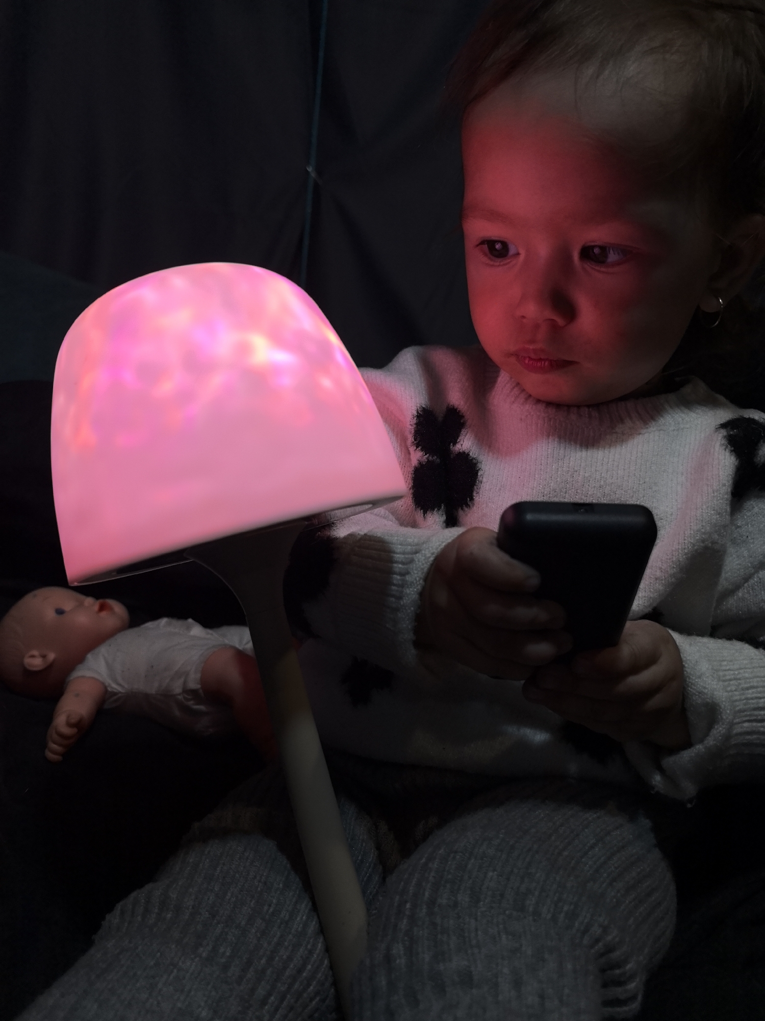 The image shows a young child seated and holding a smartphone. A mushroom-shaped lamp emitting a pink light illuminates the child's face and surroundings. A baby doll is positioned near the base of the lamp. The child wears a light-colored sweater with dark clover designs and gray pants. The setting is indoors, with a dark background. There is no clear evidence in the image to identify the location as Noviy, Russia.
