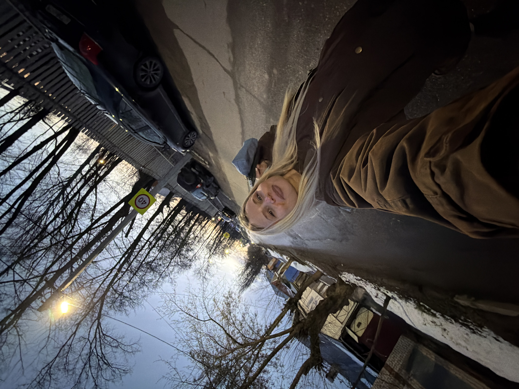 A woman with blond hair is standing on a street in Sarapul, Russia, facing the camera. She is wearing a brown jacket. Behind her is a street with parked cars, bare trees, streetlights, and buildings. A speed limit sign is visible. There is snow on the ground.