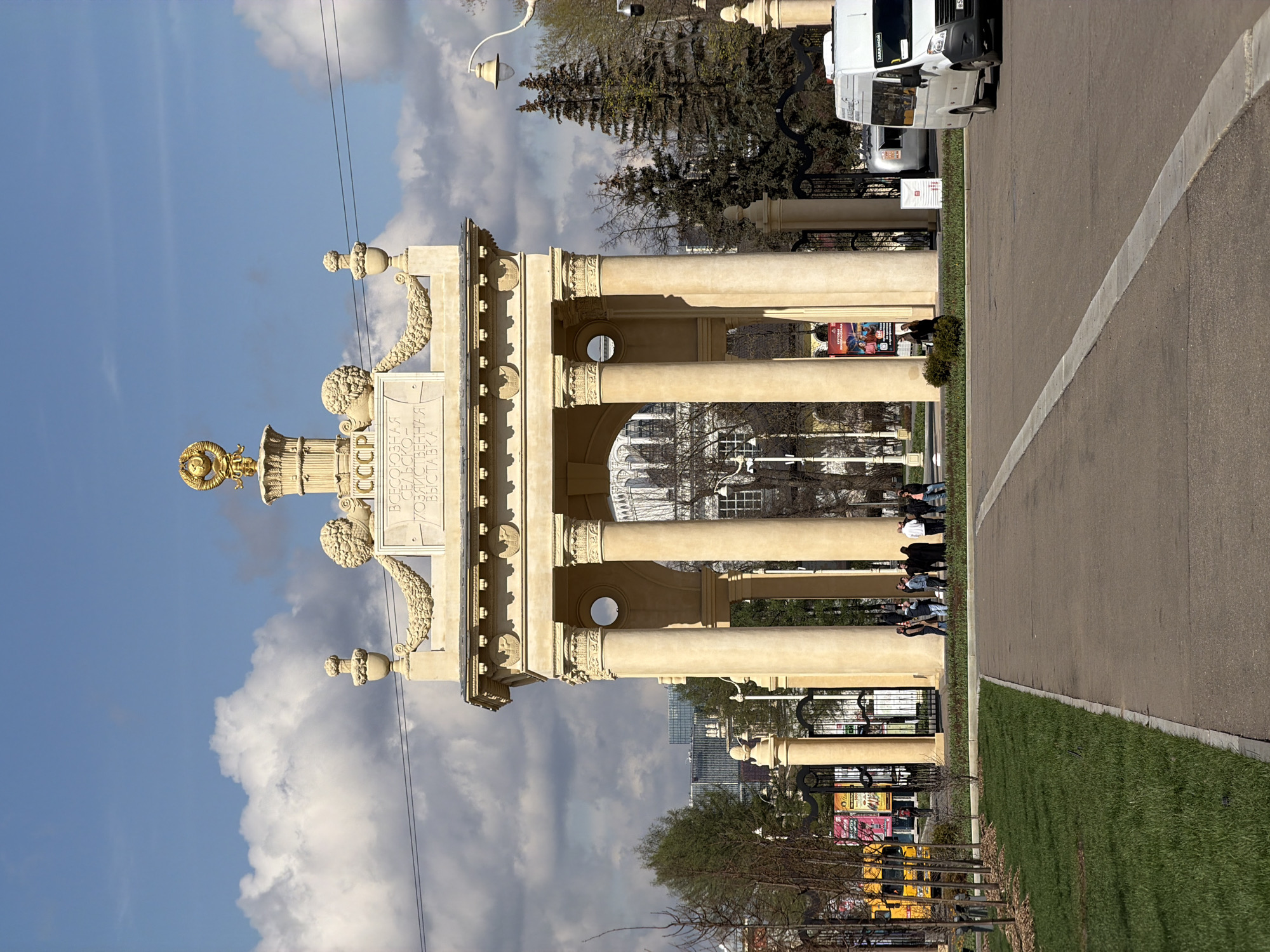 The image captures a grand, ornate architectural entrance gate or archway, characteristic of Soviet-era design. The structure is constructed from light-colored stone or concrete, featuring multiple tall, classical columns with detailed capitals and bases. Adorning the top of the archway are intricate decorative elements, including a golden emblem resembling the Soviet coat of arms (hammer, sickle, and star within a wreath) and sculptural garlands. Below this, prominent Cyrillic text is visible, reading "СССР" (USSR) and "ВСЕСОЮЗНАЯ СЕЛЬСКОХОЗЯЙСТВЕННАЯ ВЫСТАВКА" (All-Union Agricultural Exhibition), which identifies the location as VDNKh (Exhibition of Achievements of National Economy) in Moscow, Russia.
The scene takes place outdoors on a bright, sunny day with a clear blue sky dotted with scattered white clouds. To the right of the archway, a paved road with a distinct white line runs alongside a neatly maintained strip of vibrant green grass. A white commercial van is visible on this road. Through and around the archway, several people are present, some walking and others standing, suggesting a public space or attraction. Various trees, ranging from bare branches to those with emerging green leaves, are scattered in the background and along the pathways. A streetlamp is also visible in the upper left portion of the image. The overall atmosphere conveys a calm and leisurely day at a historical and cultural site.
