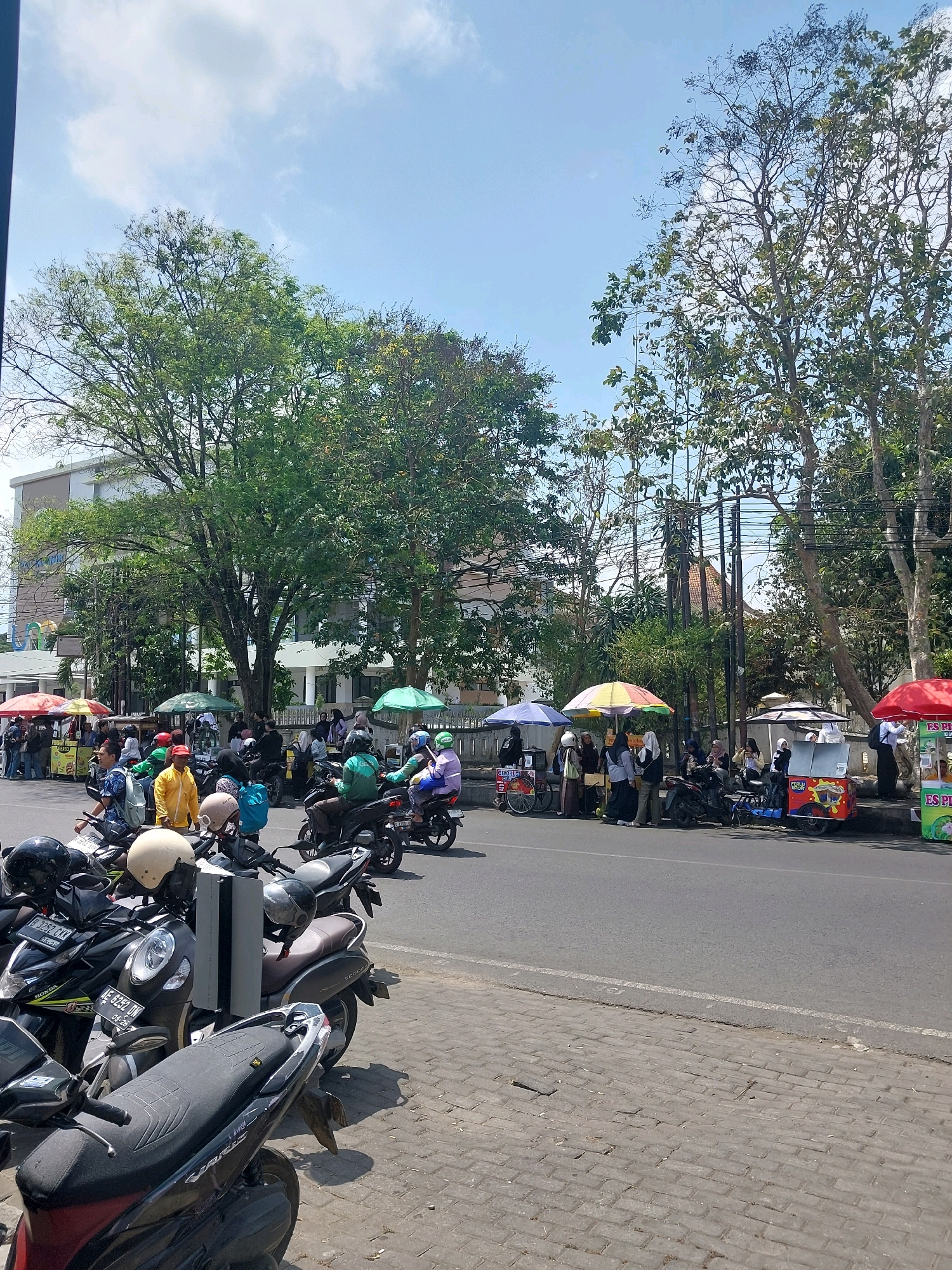 In Malang, Indonesia, a street scene unfolds under a bright, partly cloudy sky. The foreground is dominated by a row of parked motorcycles, their seats and handlebars visible. Beyond the motorcycles, a street separates the viewer from a busy sidewalk.
On the sidewalk, several food carts are set up, providing shade with colorful umbrellas. People are gathered around these carts, some standing and some sitting on their motorcycles, suggesting they are customers or vendors. A variety of people, including men and women, are present, many wearing casual clothing. Some individuals are interacting with the food carts, while others are observed in conversation or simply waiting.
Behind the sidewalk and food carts, a building with a light-colored facade stands tall, partially obscured by lush green trees. More trees line the street, their branches reaching towards the sky. The road itself appears to be an asphalt surface, with a white line marking the edge of the sidewalk. The overall impression is that of a lively, everyday scene in an urban environment, likely a popular spot for street food and daily activities.