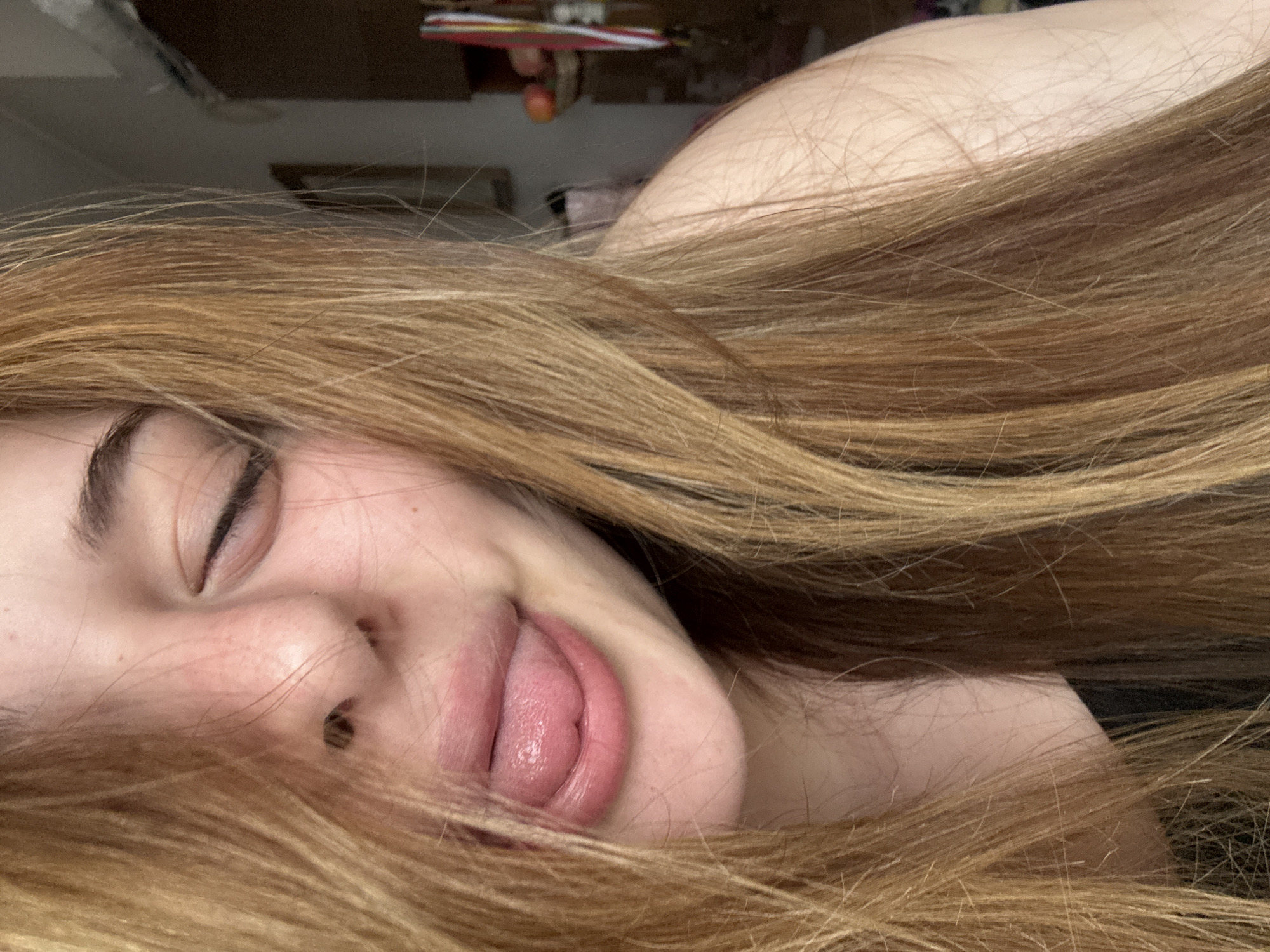 This close-up portrait, taken from a sideways perspective, features a young person with light skin and long, flowing light brown or blonde hair. The individual's face is partially visible, showing one eye that is closed or nearly closed, a nose, and a mouth with the tongue slightly extended. The hair dominates much of the frame, creating a soft, warm texture in the foreground and extending into the background.
The person appears to be resting or in a state of relaxation, indicated by the closed eye and overall serene expression, possibly with a hint of playfulness due to the tongue. A portion of the shoulder or upper chest is visible on the right side, framed by the hair. The background is softly blurred, showing indistinct shapes and colors that suggest an indoor setting, possibly a room with shelves or furniture, and a few small, unidentifiable objects. There are no specific location cues within the image to indicate Troyan, Bulgaria, nor is there any visible text. The lighting is soft and even, highlighting the features of the face.