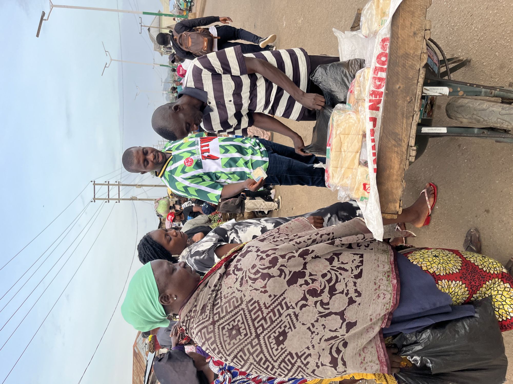 In Bukuru, Nigeria, a street vendor is seen interacting with customers. A man in a striped shirt, likely the vendor, is reaching into black plastic bags on a wooden cart filled with loaves of bread wrapped in plastic. He is facing a customer wearing a green headscarf, a patterned brown and white wrap, and a brightly colored skirt, who is looking down at the bread. Beside her, another woman in a black and white patterned dress is also observing. In the background, several other people are visible, along with market stalls and overhead electrical wires, suggesting a busy marketplace setting. The ground is paved, and the sky is overcast.