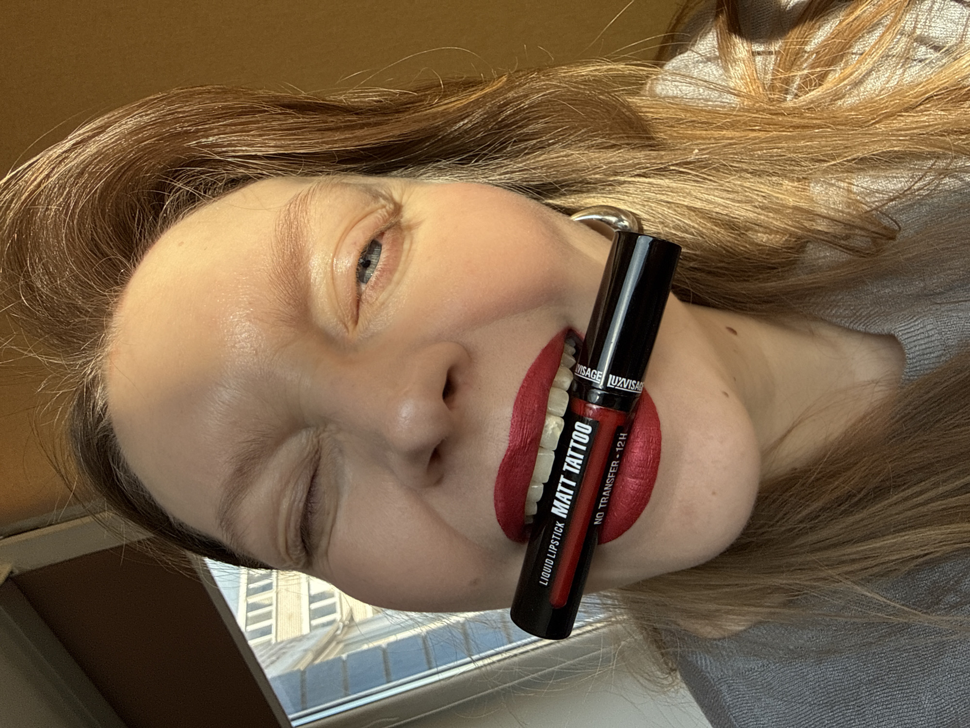 This close-up, dynamically angled portrait features an individual, likely female, with fair skin and light brown hair, captured indoors in Izhevsk, Russia. Her head is tilted, and she gazes towards the upper left with a playful or winking expression, as one eye is squinted tightly. She is showcasing a black liquid lipstick tube, held horizontally between her brightly painted, matte red lips.
The lipstick tube clearly displays white text against a red and black design. Visible inscriptions include "LUXVISAGE" (brand name at the top), "LIQUID LIPSTICK", "MATT TATTOO" (product name), and "NO TRANSFER - 12H" (a product feature or claim). The vibrant red on her lips matches the red section of the product packaging.
The scene is illuminated by bright natural light, likely from a window, creating highlights on her hair and skin and suggesting it is daytime. In the blurred background, elements of an urban setting are visible through a window, including building outlines and structures, consistent with the location context of Izhevsk. The individual is wearing a grey, possibly knitted, garment. The overall impression is a focused, product-oriented shot with a lively and engaging presentation.