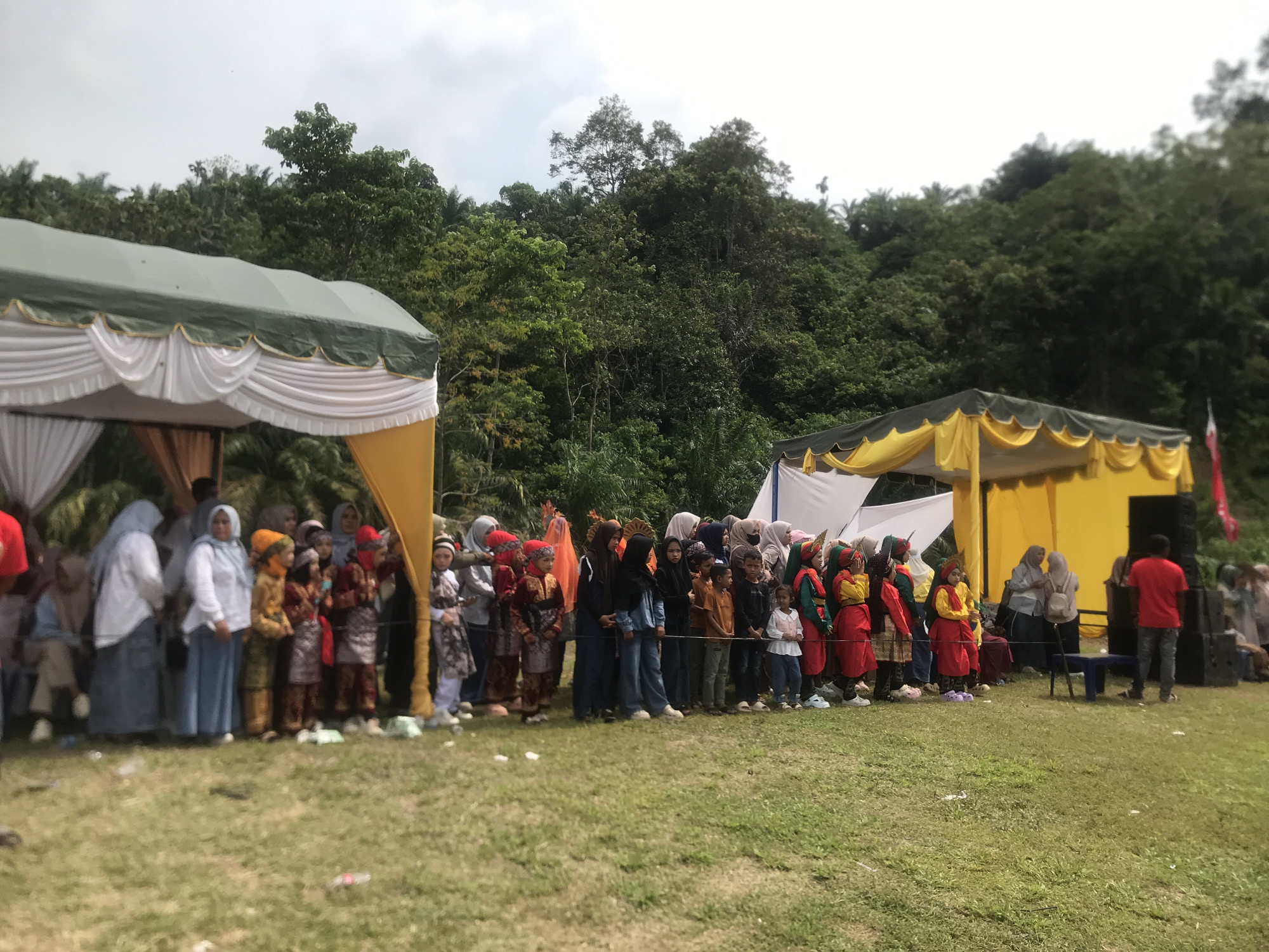 In Pantonrayeuk Satu, Indonesia, an outdoor event is taking place under a cloudy sky. Two large tents, one green with white drapes and the other yellow with white drapes, are set up on a grassy field. A line of people, including children and adults, are standing facing away from the camera, seemingly watching a performance or ceremony. Many of the individuals are wearing traditional attire, including headscarves and colorful garments. Some children are dressed in matching outfits, suggesting a group performance. In the background, a dense forest covers a hillside. To the right of the yellow tent, a black speaker system is visible, and a man in a red shirt is standing near it. The overall scene suggests a community gathering or cultural event.