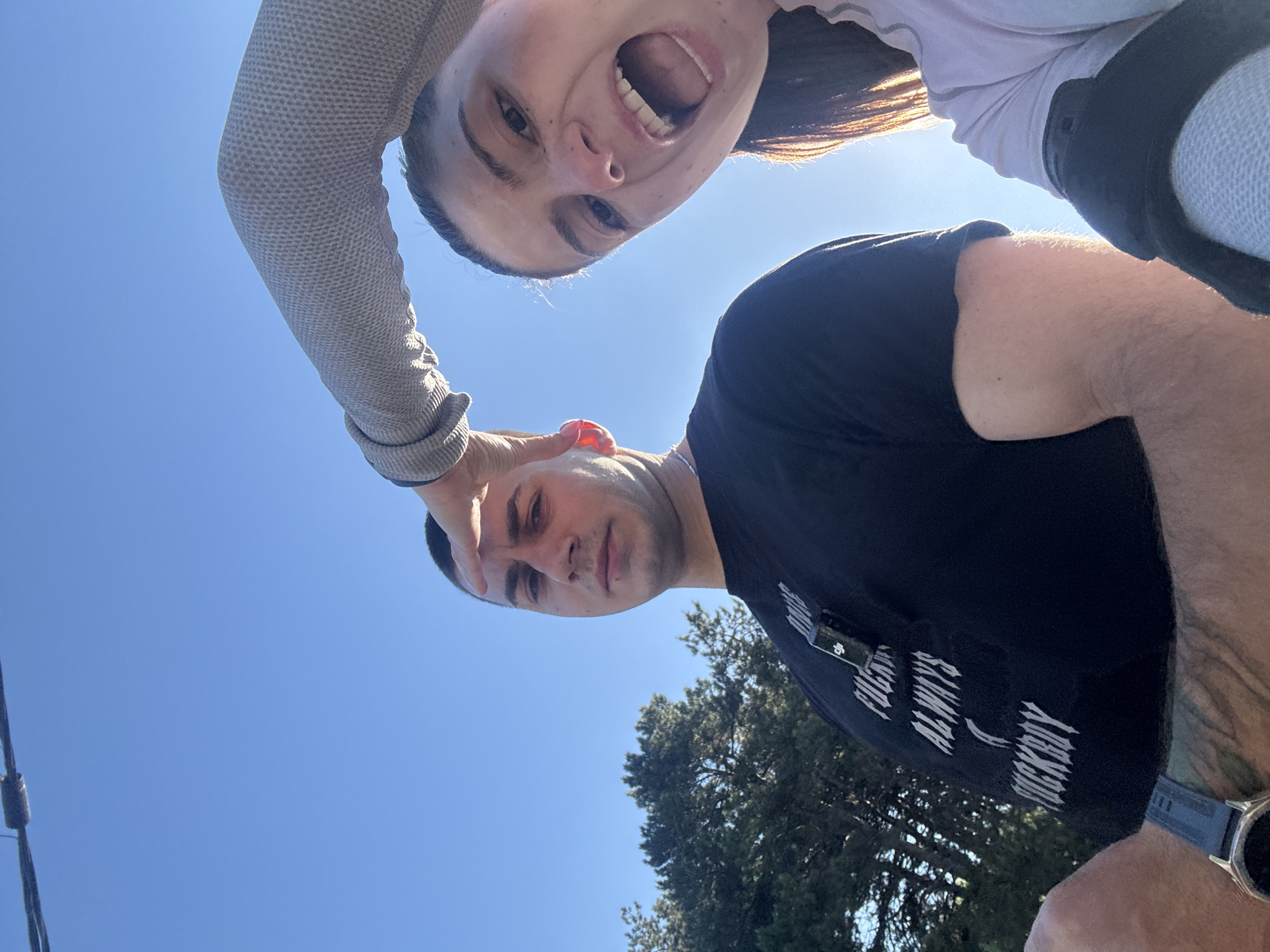 The image shows two individuals in an outdoor setting. The individual on the bottom, a man, is wearing a black t-shirt with white text. He has short hair and is looking downward. The individual on the top, a woman, is wearing a light colored top. The photo appears to have been taken from a low angle, with a clear blue sky visible in the background. There are trees in the background. There is no way to discern the location in the city of Varna, Bulgaria.
