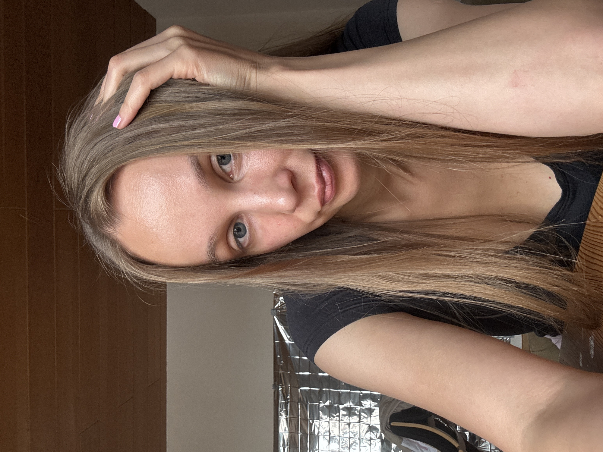 A woman with long, straight, light brown hair is leaning her head on her hand. She is looking at the camera with blue eyes and a neutral expression. She is wearing a black t-shirt. The background is out of focus, but appears to be an indoor setting with a wooden ceiling and a reflective surface behind her. There is no information about the location in the city of Überherrn, Germany.