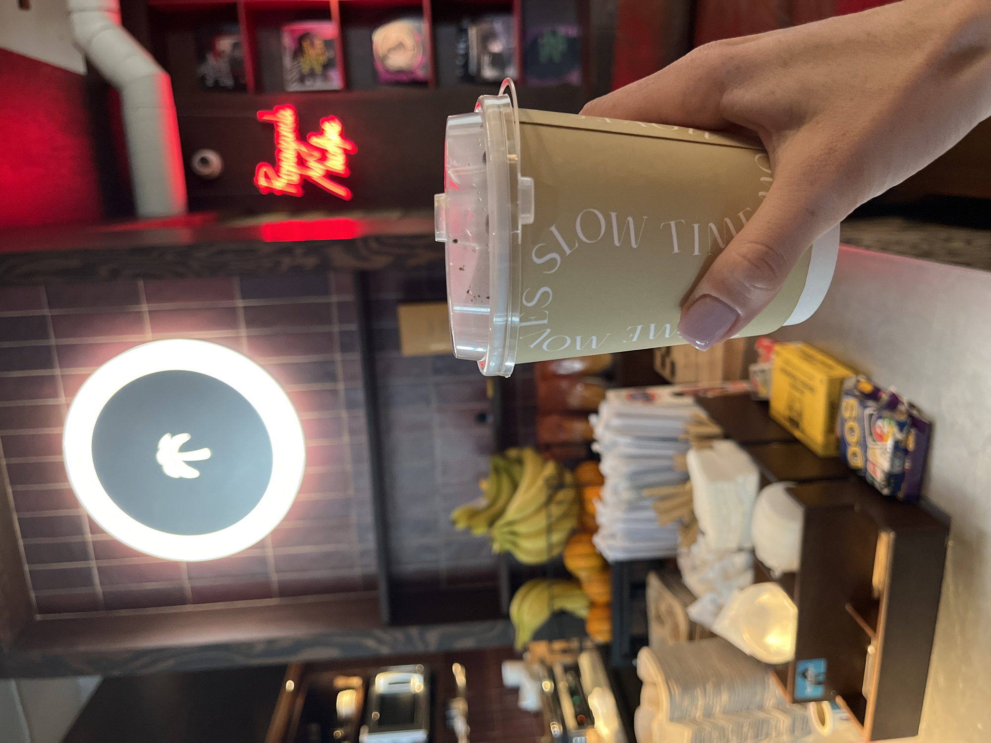 A person's hand, with light purple nail polish, holds a beige disposable coffee cup with a clear plastic lid. The cup has white text printed in a circular pattern, "SLOW TIME," visible on its side. The background suggests an indoor establishment, possibly a cafe or bar. A circular sign with a stylized palm tree in white on a dark gray background is illuminated by a bright white halo. To the right, a red neon sign reads "Propaganda Machine." Shelves in the background display various items, some appearing to be records or framed art. In the mid-ground, a counter displays stacked paper cups, napkins, and what appear to be small pumpkins and bananas. A microwave or similar appliance is visible on a shelf to the left. The scene is lit by a combination of ambient light and the neon sign, creating a warm and inviting atmosphere. This establishment is located in Kazan, Russia.