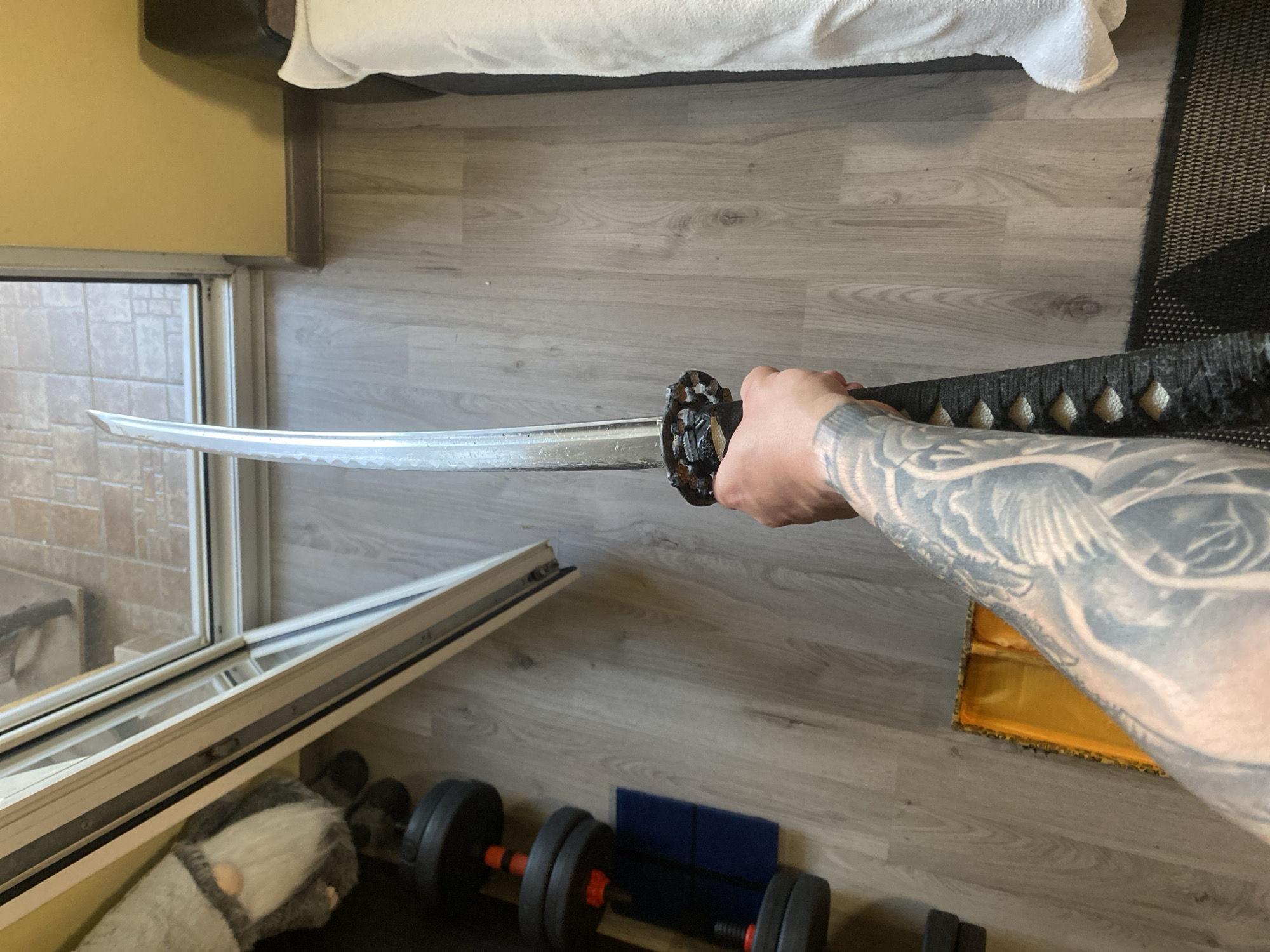 A male individual is depicted holding a katana-style sword vertically by its handle in an interior residential setting in Varna, Bulgaria. The individual's left forearm and hand are visible, revealing extensive black and grey tattoo work, possibly depicting a mythological creature. The sword features an ornate, dark-colored circular tsuba and a black-wrapped tsuka. The blade is partially visible, showing a polished metallic surface.
The floor is covered with light-toned laminate wood planks. To the left, a set of black adjustable dumbbells with red accents rests on the floor, positioned against a dark surface. Adjacent to the dumbbells is a decorative grey gnome figure with a white beard. Further left, a large, white-framed sliding glass door provides a view to an exterior tiled patio area and a distant reddish-brown brick structure. A piece of yellow or gold fabric is visible on the floor near the lower left quadrant.
To the right, a pale yellow-painted wall is visible. A portion of a bed or couch, covered with a white textured blanket or sheet, occupies the right side of the frame. A dark headboard or frame is partially visible behind the bedding. A black, textured rug is situated on the floor in the lower right corner, extending beneath the bedding. The room is illuminated by natural light entering through the glass door.