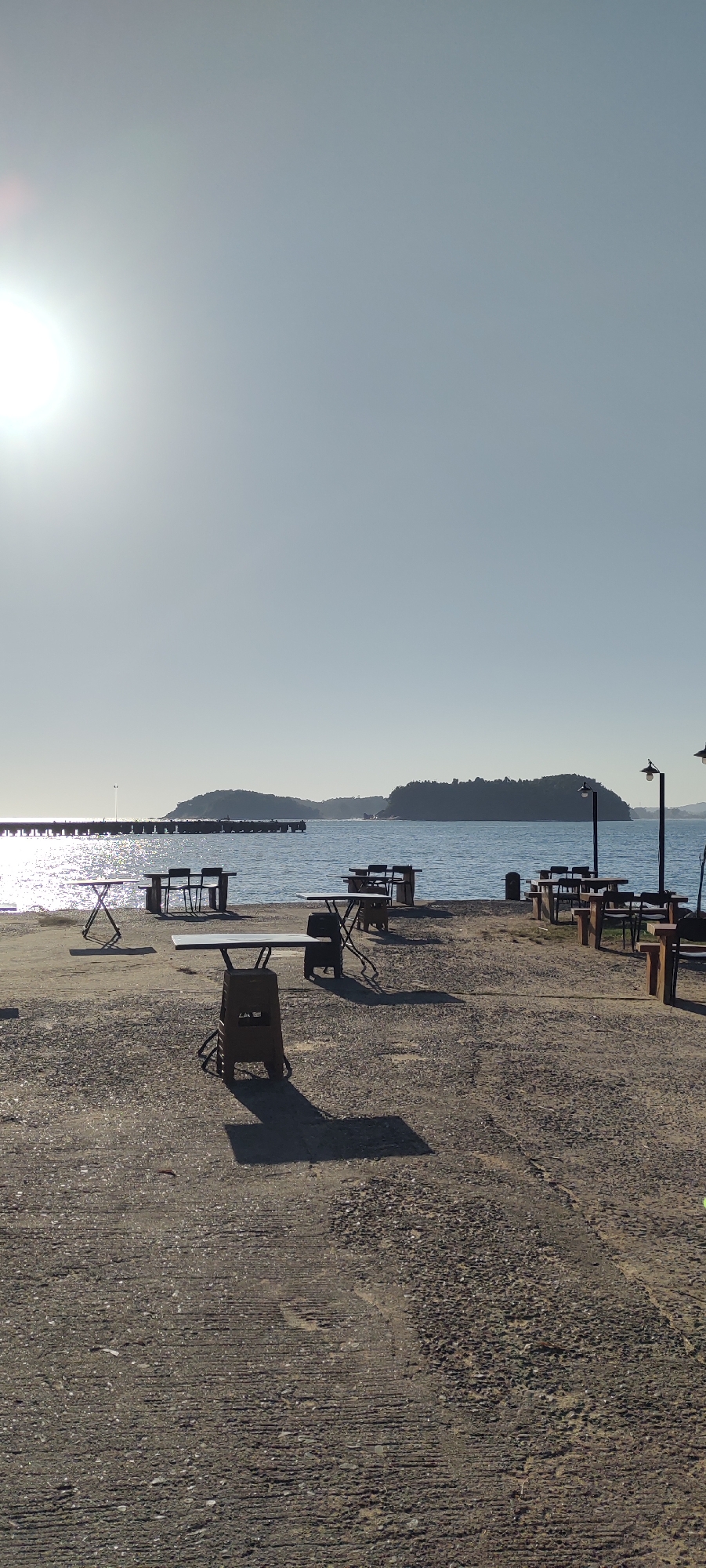 The image shows an outdoor seating area near a body of water, likely the sea, under a clear, bright sky with the sun prominently featured in the upper left quadrant. The foreground is a concrete ground surface with scattered gravel and visible tire marks. Several empty tables and chairs are set up, some made of metal with a distressed finish, others appearing to be plastic. The tables are of various designs, including folding tables and sturdier ones supported by stacked plastic crates. Long shadows are cast by the tables and chairs, indicating a sunny day.
In the middle ground, the water stretches out, reflecting the sunlight. A long pier or jetty extends into the water from the left side of the frame. On the right side, there are two street lamps with downward-facing lights. In the distance, across the water, is a hilly, tree-covered island.
There are no people visible in the scene. The overall atmosphere is quiet and undisturbed, suggesting a place that is either closed for the day or not yet open for business. The location in Calang, Indonesia, is not explicitly identified by any signage or landmarks within the image itself, but the description of a coastal setting with islands and a pier aligns with the geographical characteristics of coastal Indonesia.