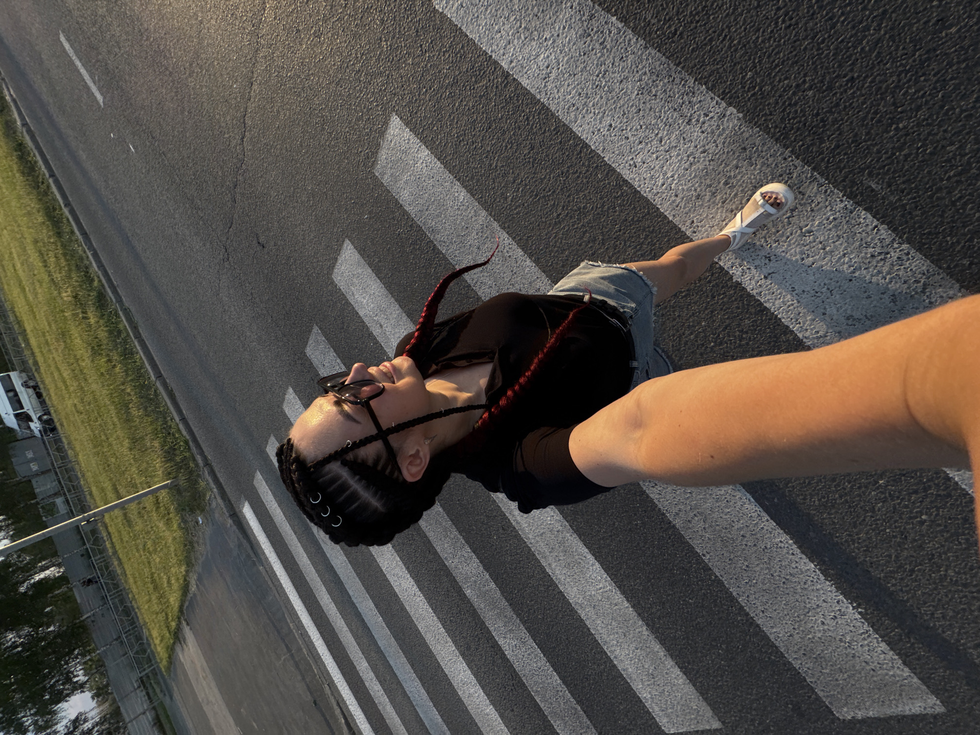 In a high-angle, full-body shot, a young woman with braided hair stands on a crosswalk. She is wearing sunglasses, a black t-shirt, and denim shorts, with white sandals on her feet. Her right arm is extended forward, holding a camera as she takes a selfie. The background shows a grassy area with a fence and a white van parked in the distance, suggesting an outdoor urban setting during daylight. The lighting indicates a warm, late afternoon sun.