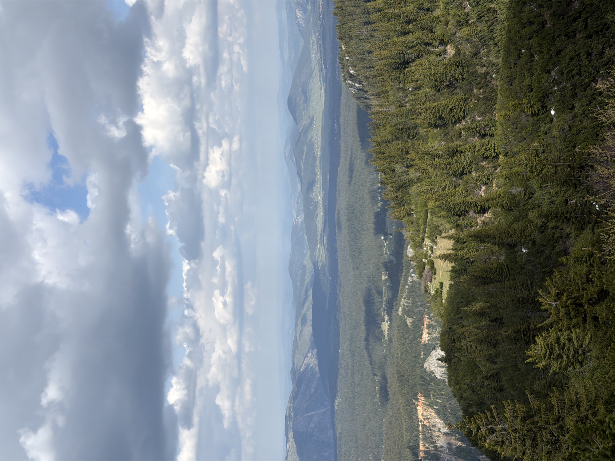The image presents a landscape view from an elevated position in Bosača, Montenegro. The sky on the left exhibits variable cloud cover, transitioning from dark, heavy clouds to patches of blue sky. The landscape comprises layers of mountain ranges receding into the distance. Predominantly, the terrain is covered in dense forest vegetation. Evidence of exposed rock formations or cliffs are visible in the lower right section.