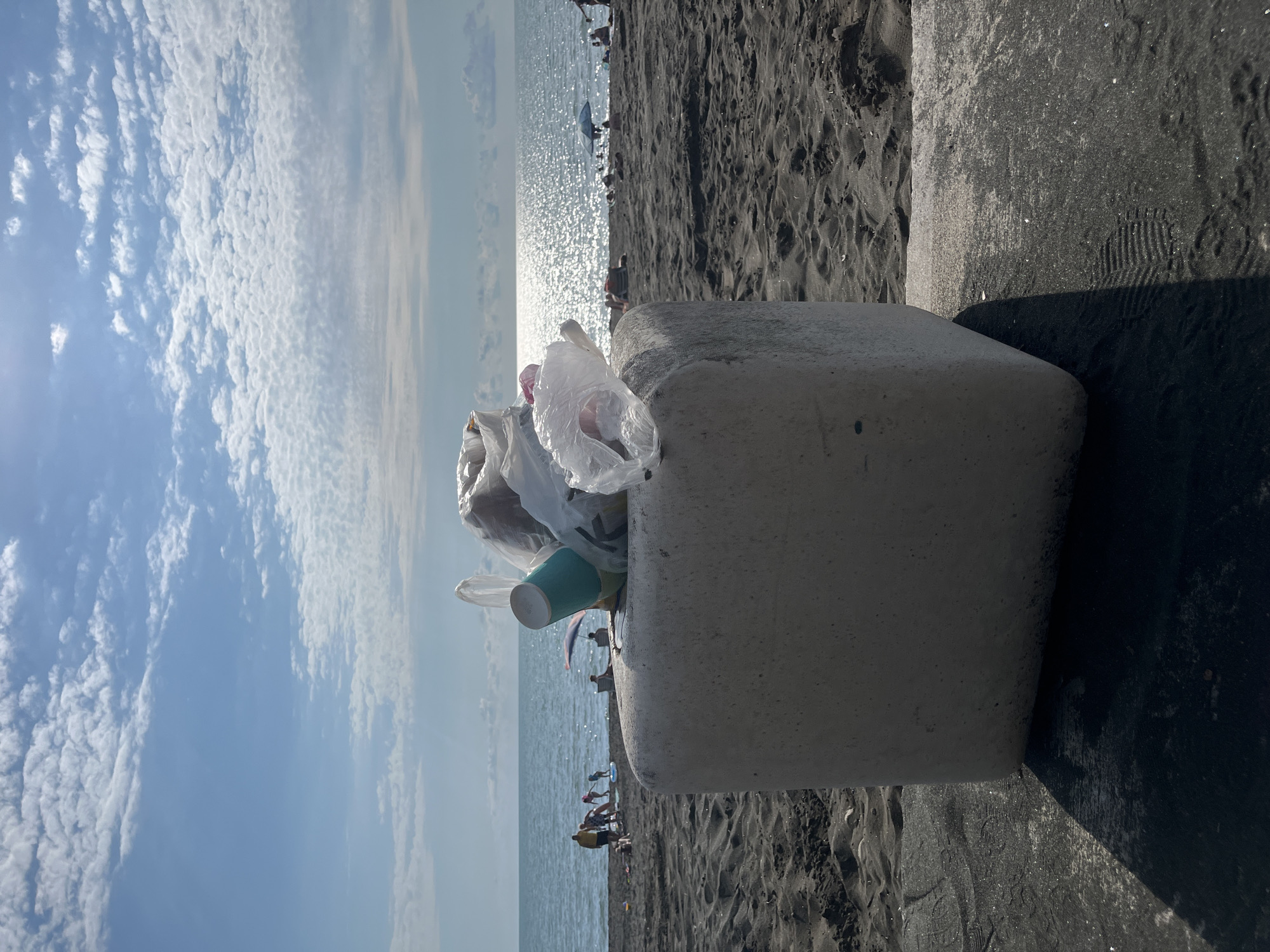 This media file captures a scene on a beach in Ureki, Georgia. The primary subjects are a concrete trash receptacle overflowing with plastic bags and a single blue cup, and the surrounding environment. The setting is a beach with dark sand, the calm sea, and a sky filled with scattered white clouds.
In the background, several people are visible, some near the water's edge and others further back on the sand. They appear to be engaged in leisurely beach activities, though their specific actions are not clearly discernible due to the distance and resolution. The sun is shining brightly, reflecting off the water and casting shadows, suggesting it is daytime, likely late morning or early afternoon. The overall mood conveyed is one of a typical beach day, albeit with a prominent element of litter.
Notable details include the abundance of plastic waste in the trash bin, which stands out against the natural backdrop of the beach and sea. The concrete structure of the bin is rough and weathered. The sand has a fine, dark texture, and there are footprints visible in it, indicating recent human activity. The sea is a deep blue, with gentle waves. The sky is predominantly blue with soft, wispy clouds that are illuminated by the sun. No visible text is present in the media file. The image is oriented vertically.