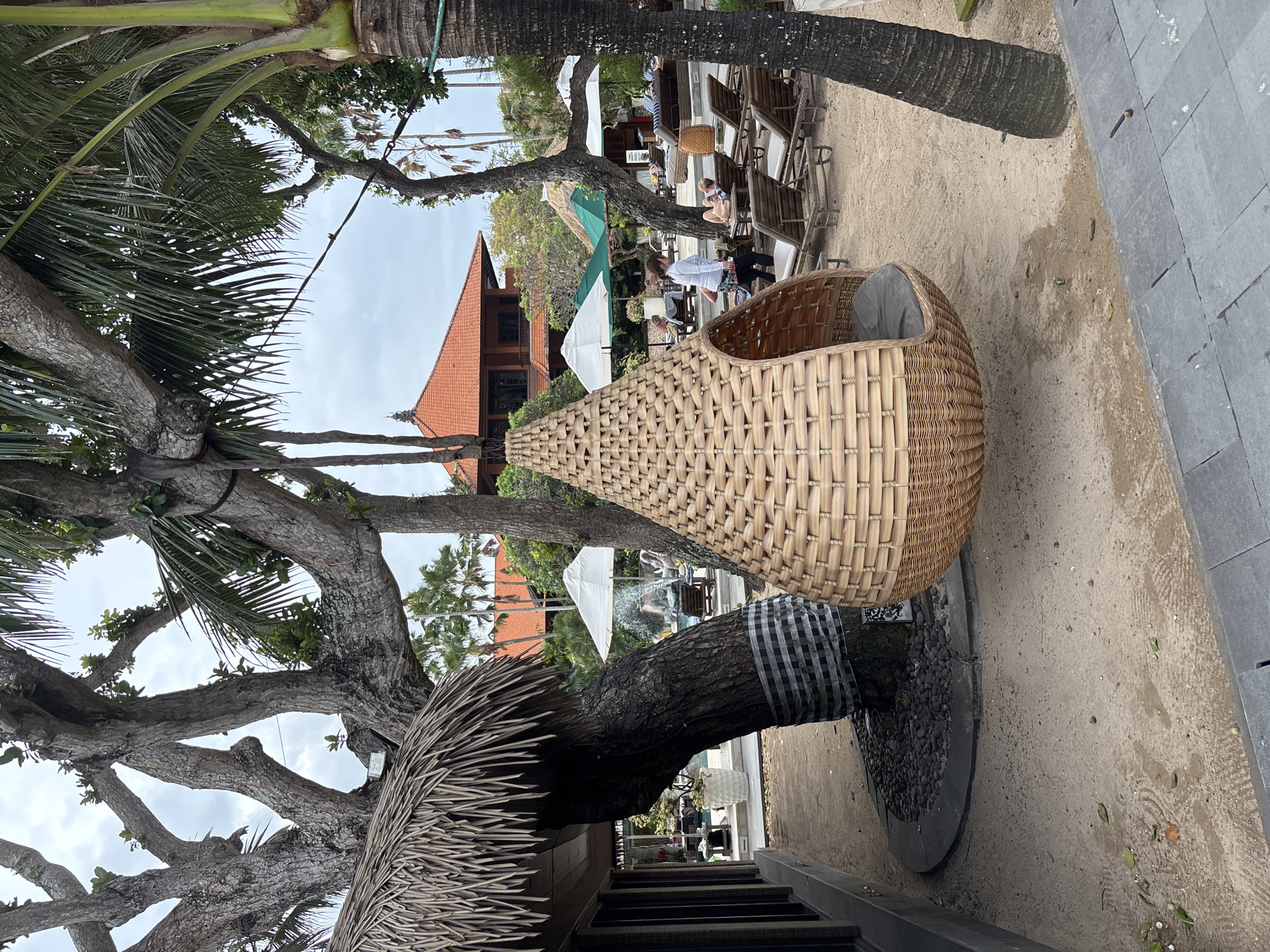 A hanging woven egg-shaped chair with a gray cushion is suspended from a tree branch on a sandy beach in Denpasar, Indonesia. The chair is made of light brown wicker and has an opening on one side. In the background, there are several lounge chairs, umbrellas, a swimming pool, and a building with an orange tiled roof. Palm trees and other tropical foliage are visible throughout the scene. A person is walking away from the camera towards the lounge chairs, and other people are visible relaxing on the chairs in the background. The sky is overcast.