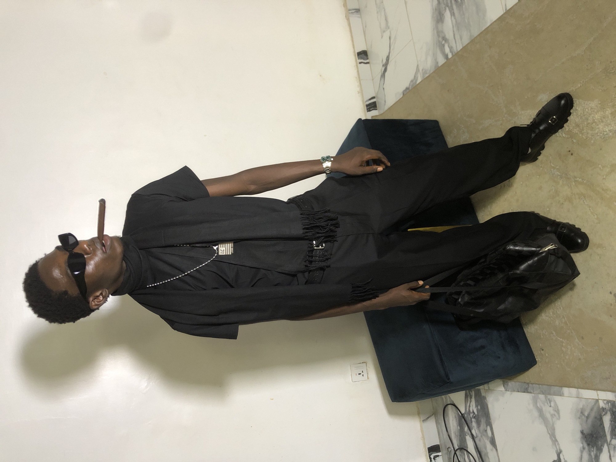 A young Black male stands in a room, dressed entirely in black clothing. He wears black sunglasses, a black turtleneck, a black scarf with fringed ends, and black pants. A silver chain with a pendant is visible around his neck. He has a cigar held between his lips and is holding a black duffel bag in his left hand. A watch is on his right wrist. The background consists of a white wall, a dark blue upholstered bench, and a marble-patterned floor. The scene appears to be indoors, possibly in a residential setting, and does not provide specific indicators of being in the city of Ado, Nigeria.